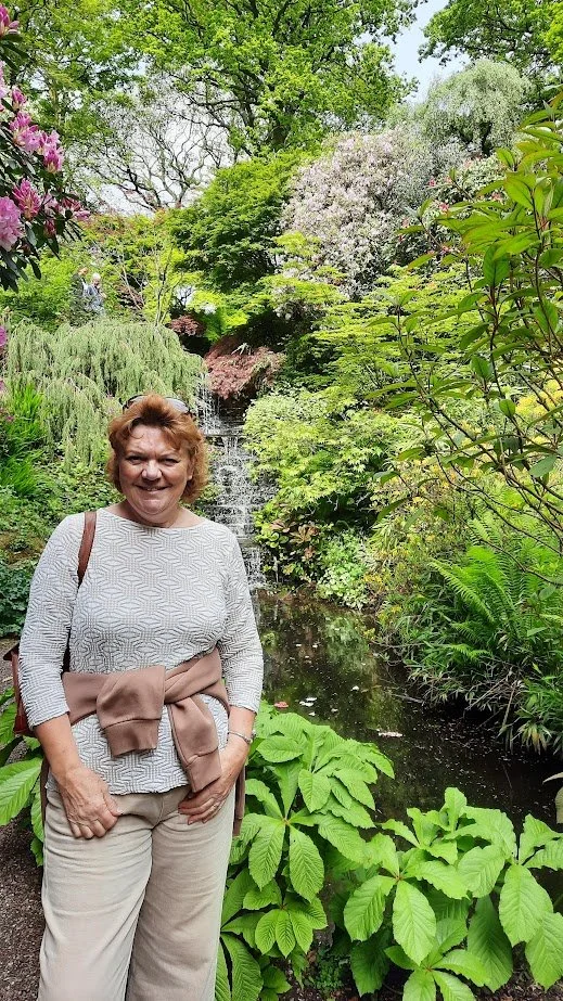 A woman smiling in a lush green garden with a waterfall in the background, surrounded by various plants and trees.