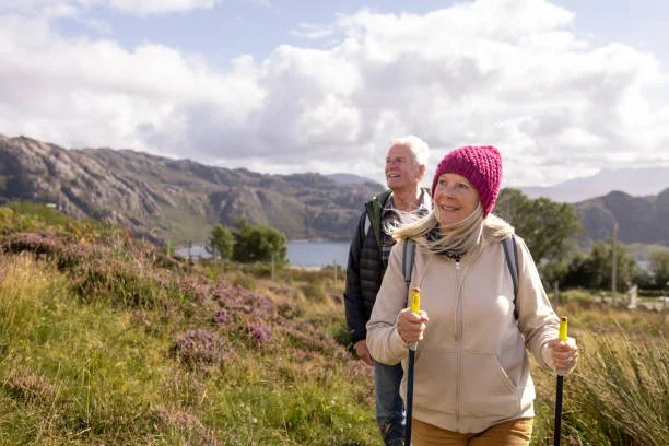 Older couple hiking on a trail near a lake with mountains in the background, woman wearing a pink knit hat and holding trekking poles, man wearing a dark jacket behind her.