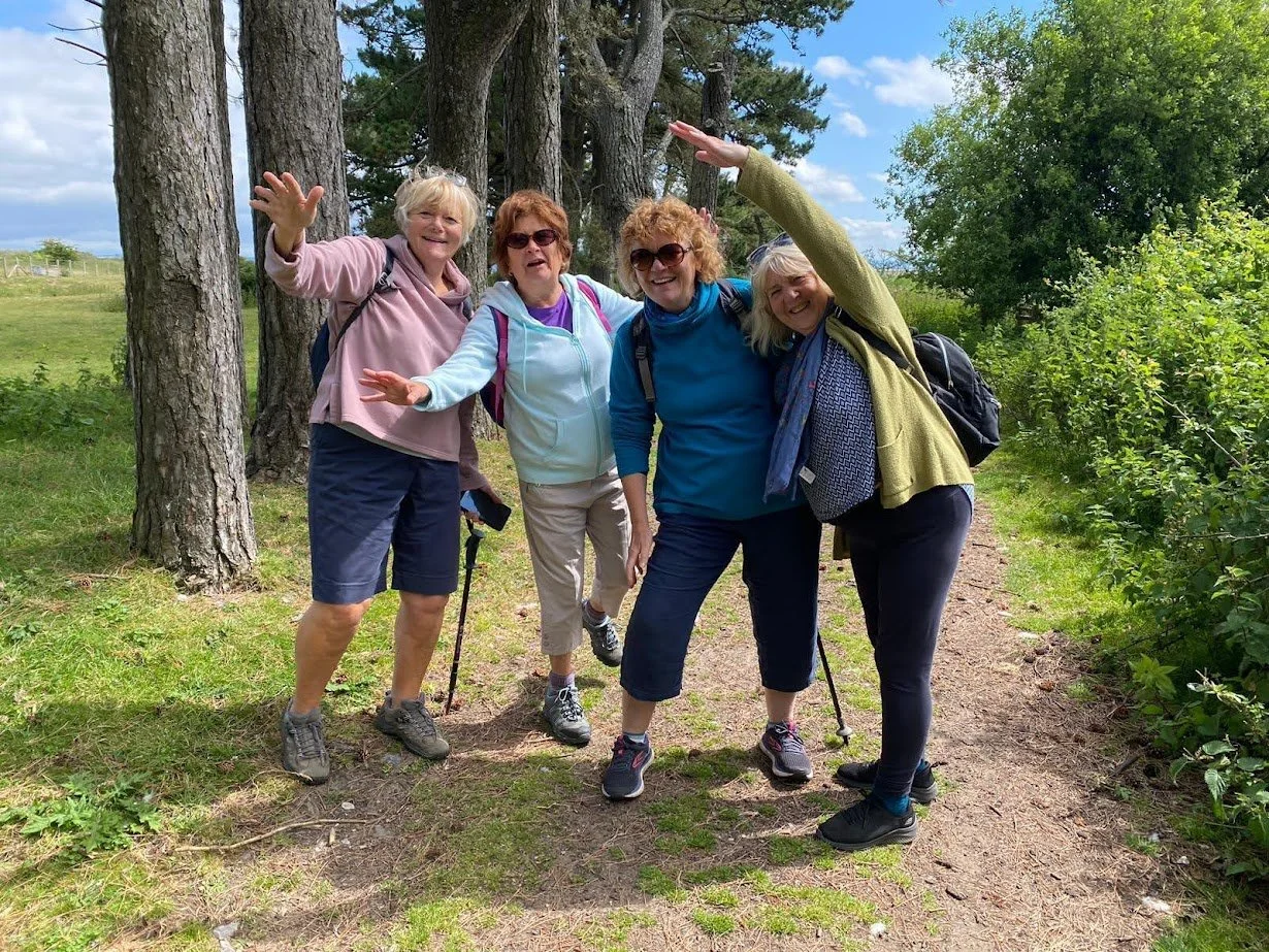 Four women posing happily on a forest trail surrounded by trees and greenery.