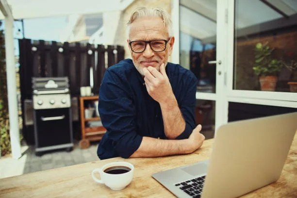 An elderly man with glasses and gray hair, sitting at a wooden table outside, looking at a laptop with a thoughtful expression, with a cup of coffee next to him.