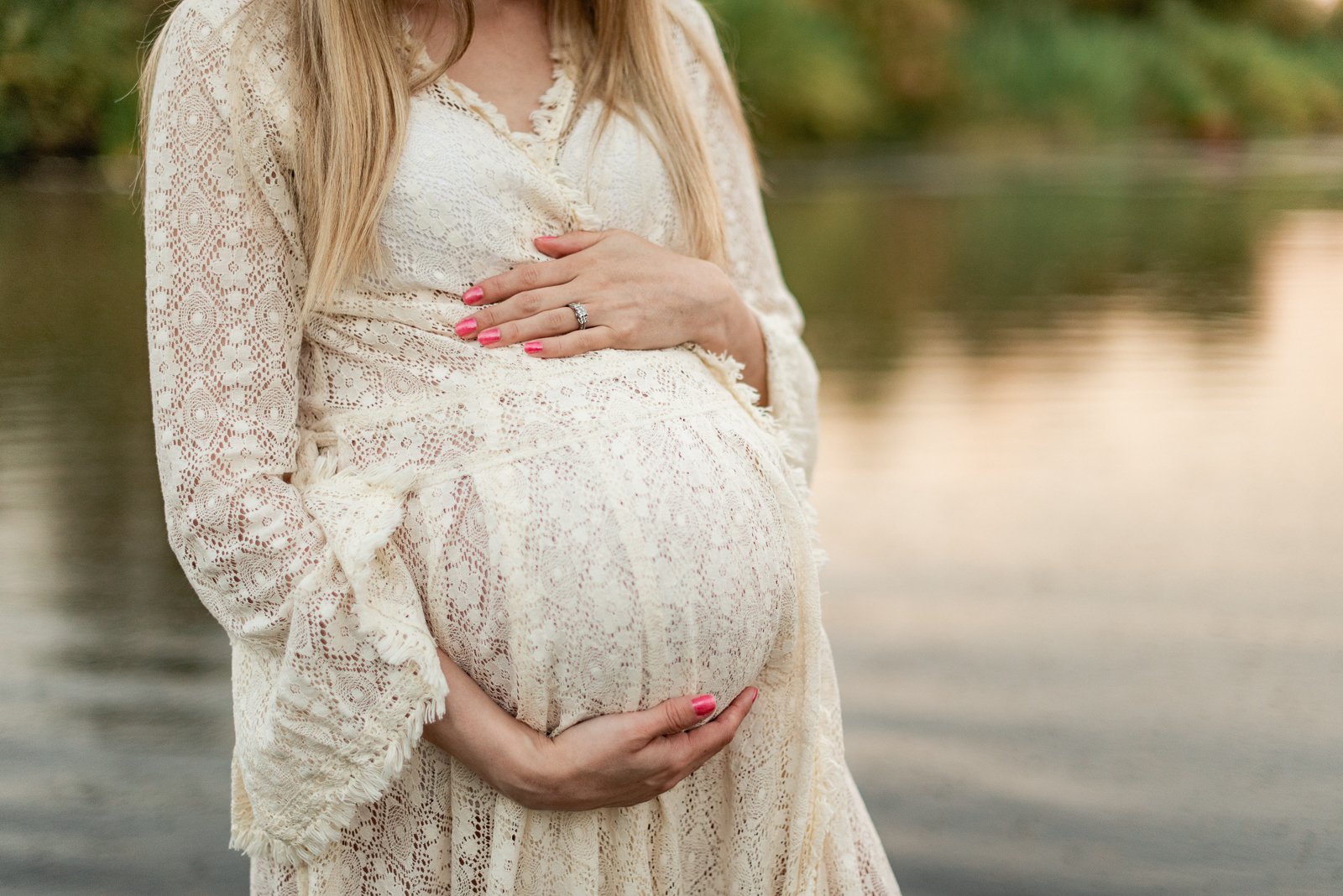 A pregnant woman wearing a cream-colored lace dress, holding her baby bump with both hands, standing near a body of water outdoors.