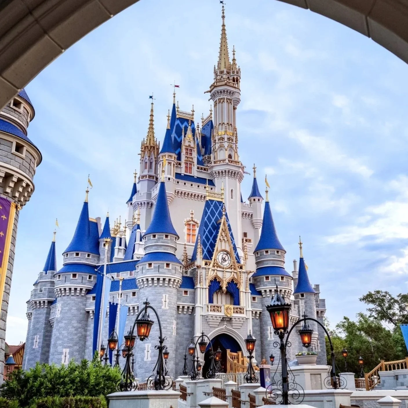 Disney Castle with blue roofs and gold accents, viewed through an archway, under a partly cloudy sky.