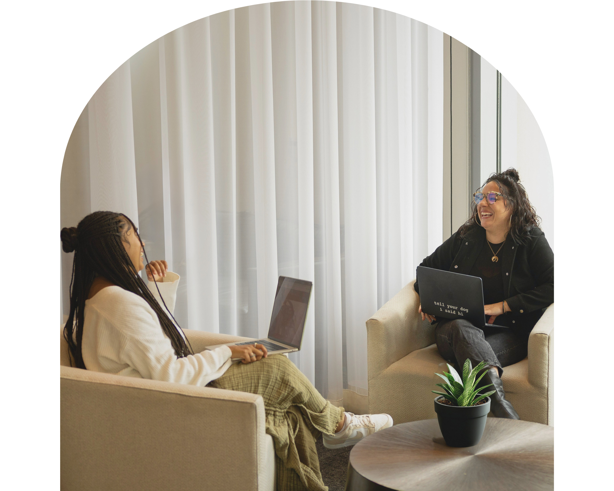 Two women having a conversation in a modern, well-lit room with white curtains. One woman is sitting on a beige couch with a laptop, wearing a white sweater and checkered pants. The other woman is sitting on an armchair, wearing glasses and a black jacket, holding a small blackboard that says 'tell your dog I said hi.' There is a potted plant on a small round table in the foreground.