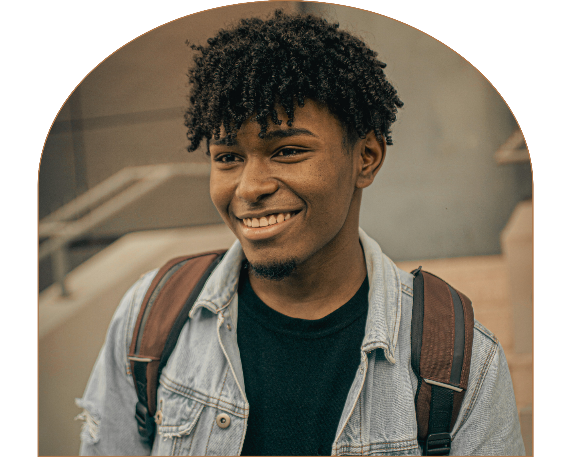 A young person with curly black hair, smiling, wearing a denim jacket over a black shirt, and carrying a brown backpack.