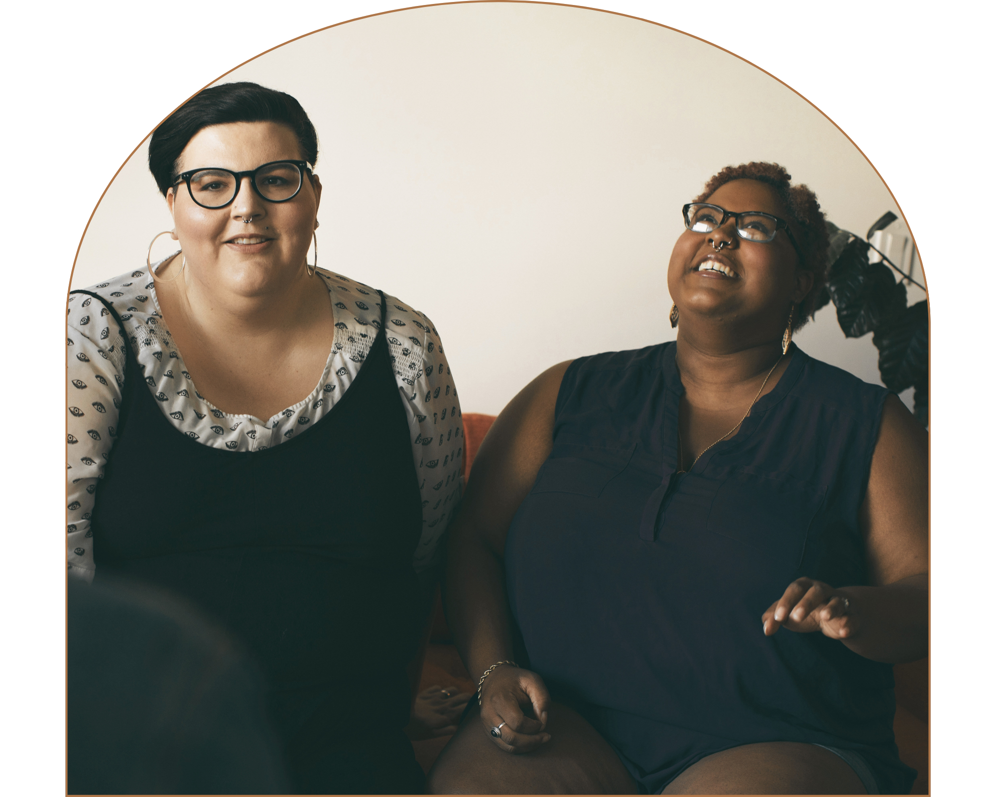 A couple sitting close together, one with short dark hair, glasses, and a nose ring, smiling at the camera, and the other with curly hair, glasses, and jewelry, looking up and smiling.
