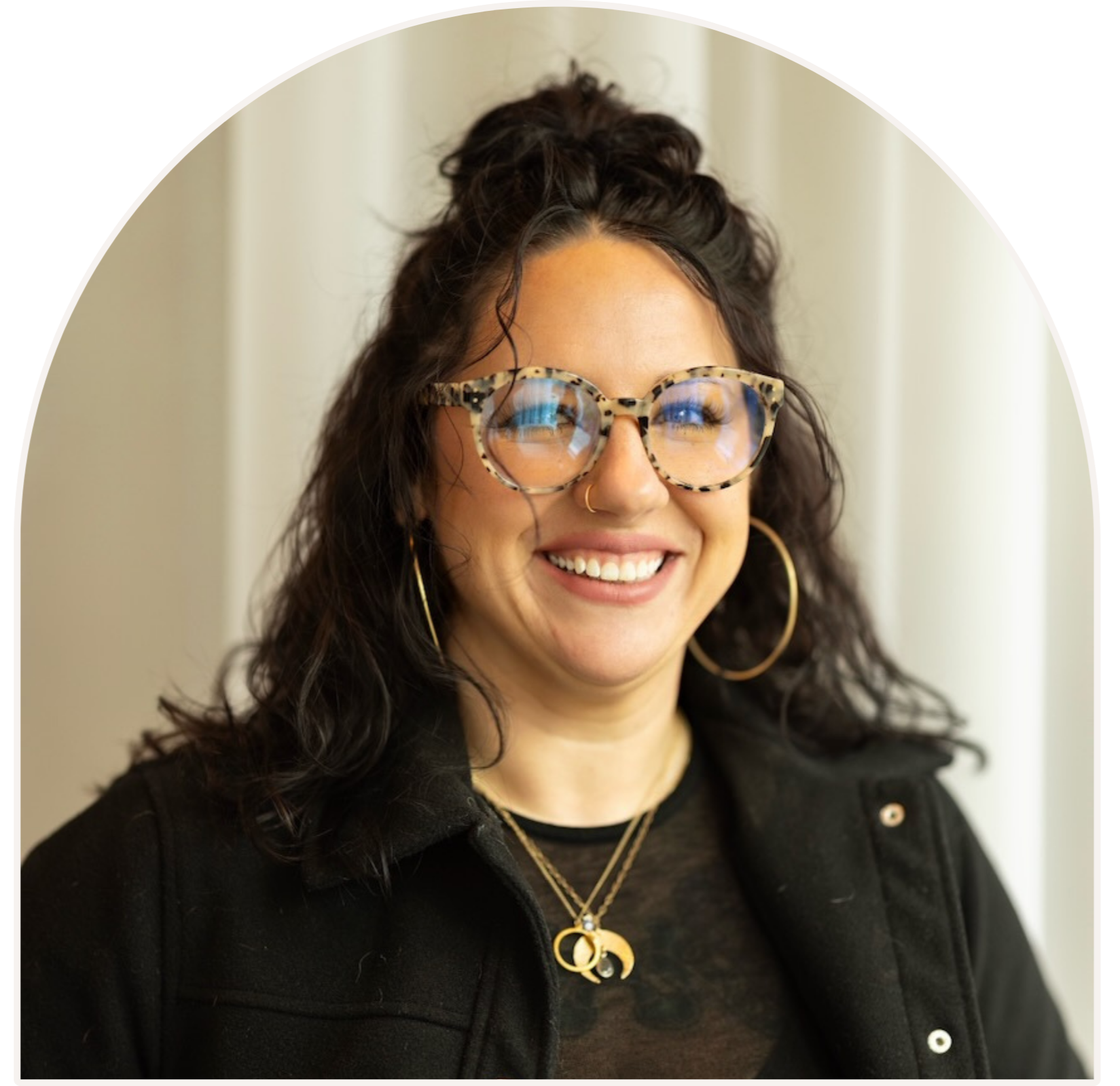 Clinician and co-owner Megan, smiling with curly dark hair, wearing large tortoiseshell glasses, gold hoop earrings, and layered necklaces, dressed in a black jacket over a black top, standing in front of a light-colored curtain.