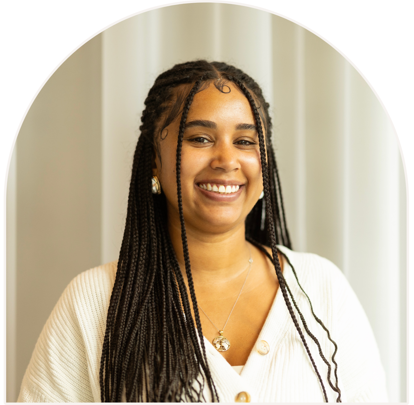 Clinician and co-owner Kiara, smiling with dark braided hair, wearing earrings, a necklace, and a cream-colored cardigan, standing in front of a light-colored curtain.