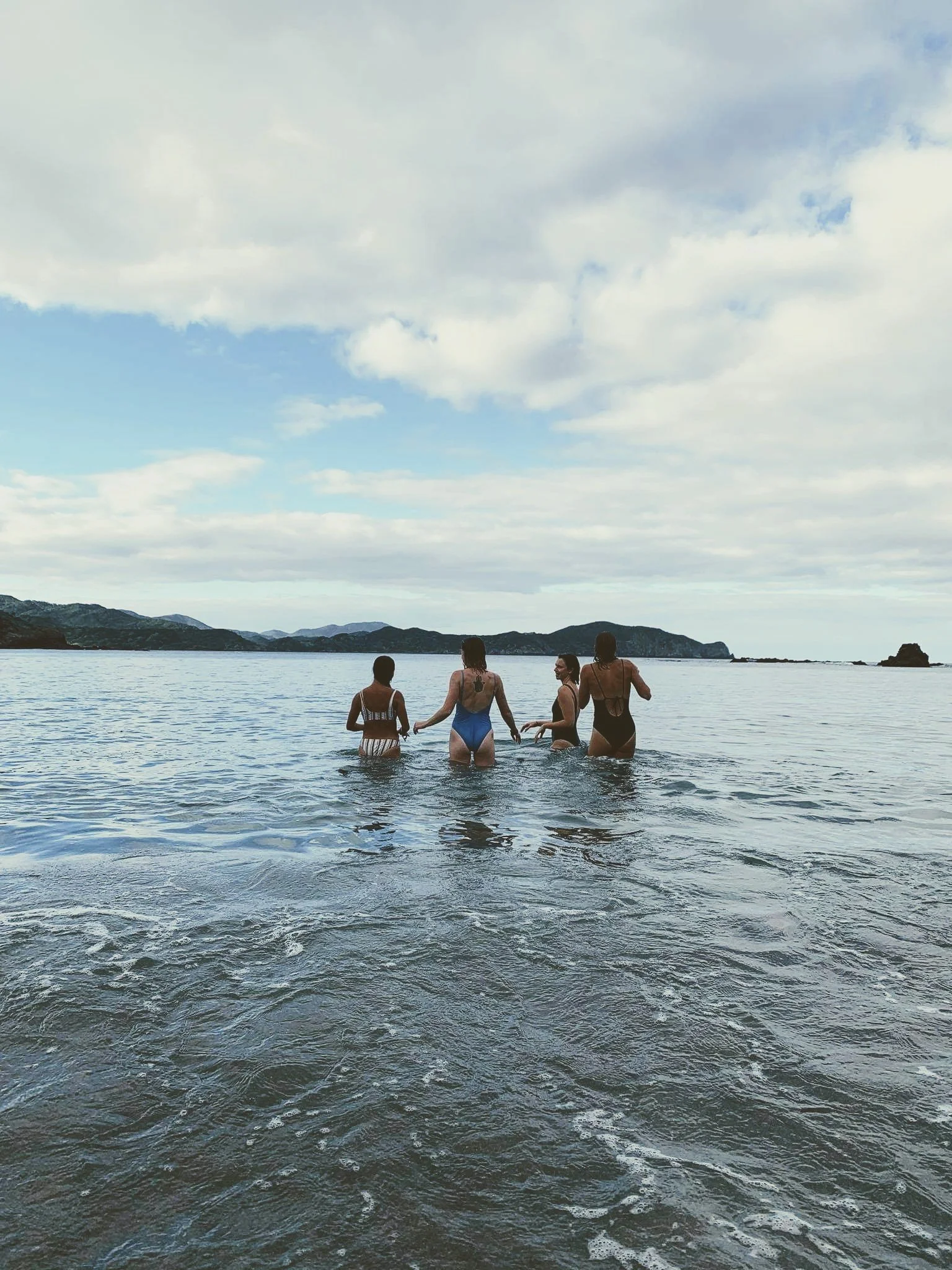 Four women in swimsuits standing waist-deep in a calm body of water, facing away from the camera beneath a cloudy sky with mountain views in the background.