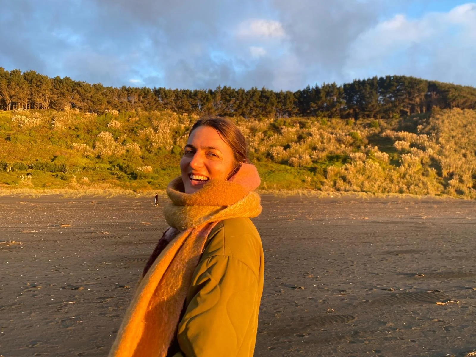 A woman with a bright smile standing on a beach during sunset, wearing a multicolored scarf and olive green jacket, with a hillside covered in trees and shrubs in the background.