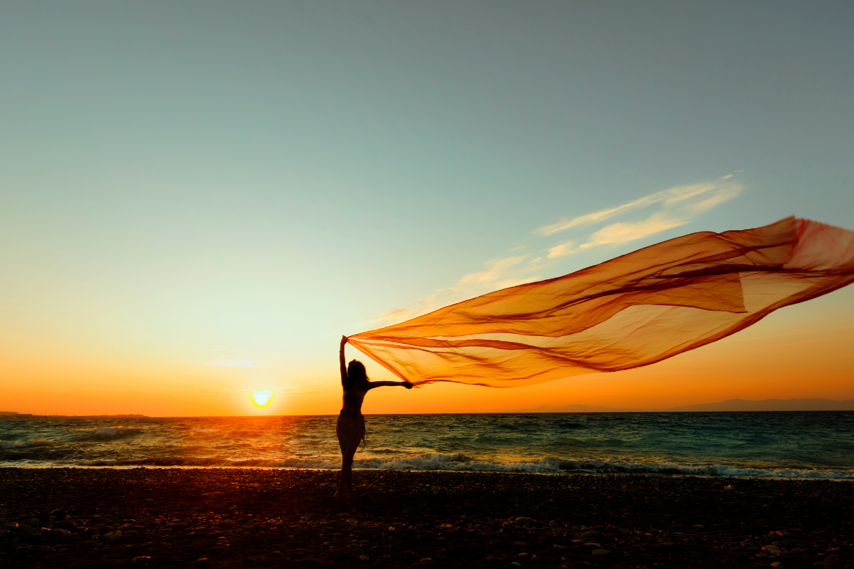A woman holding a flowing fabric on a beach during sunset.