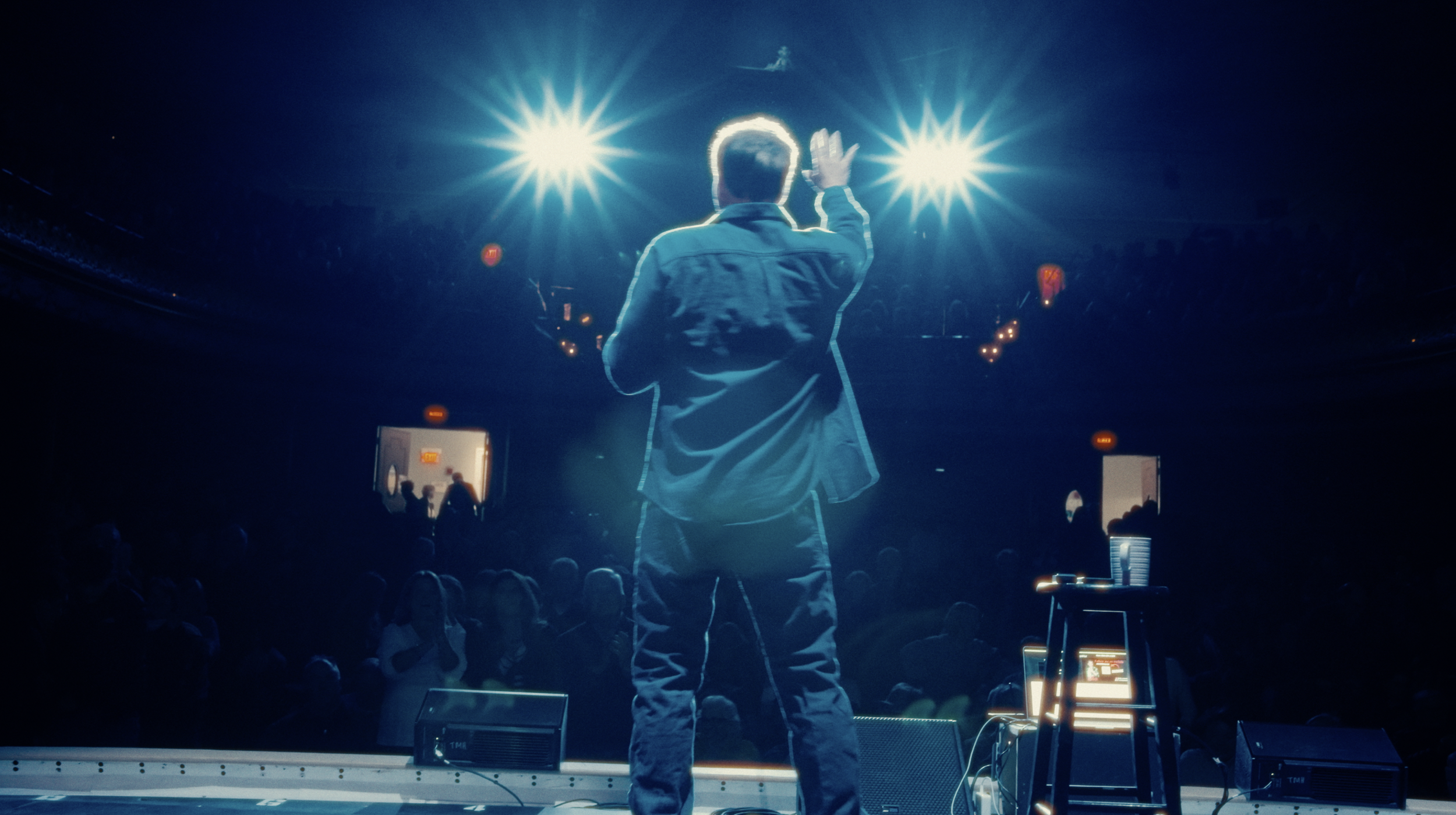 A man on stage with his back to the camera, raising his right hand as if waving, in front of bright stage lights, with an audience watching.
