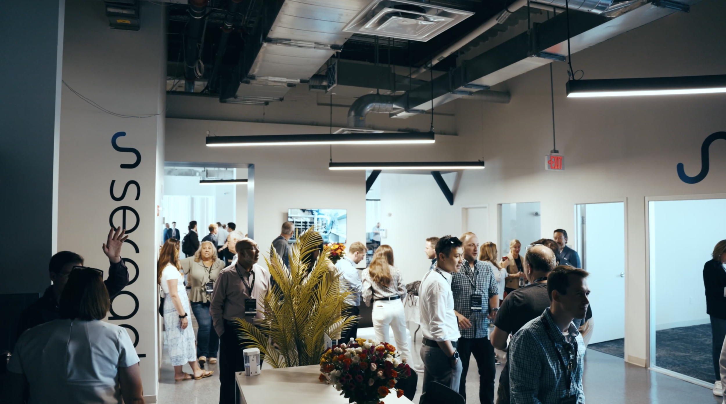 Crowd of people at a tech conference or event inside a modern building with industrial ceiling and signage. People are engaging, talking, and walking around.