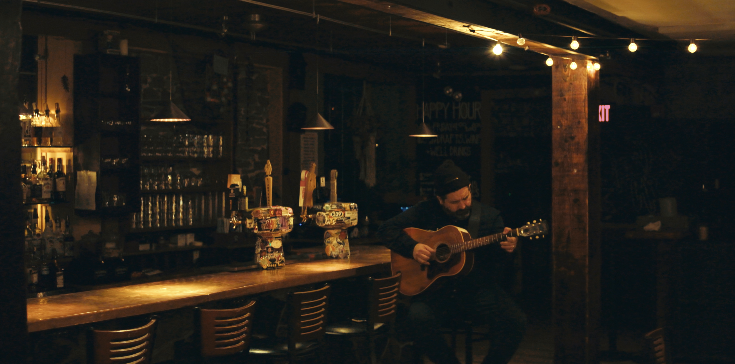 A man playing an acoustic guitar in a dimly lit bar with exposed brick walls, liquor bottles on shelves, and warm lighting.