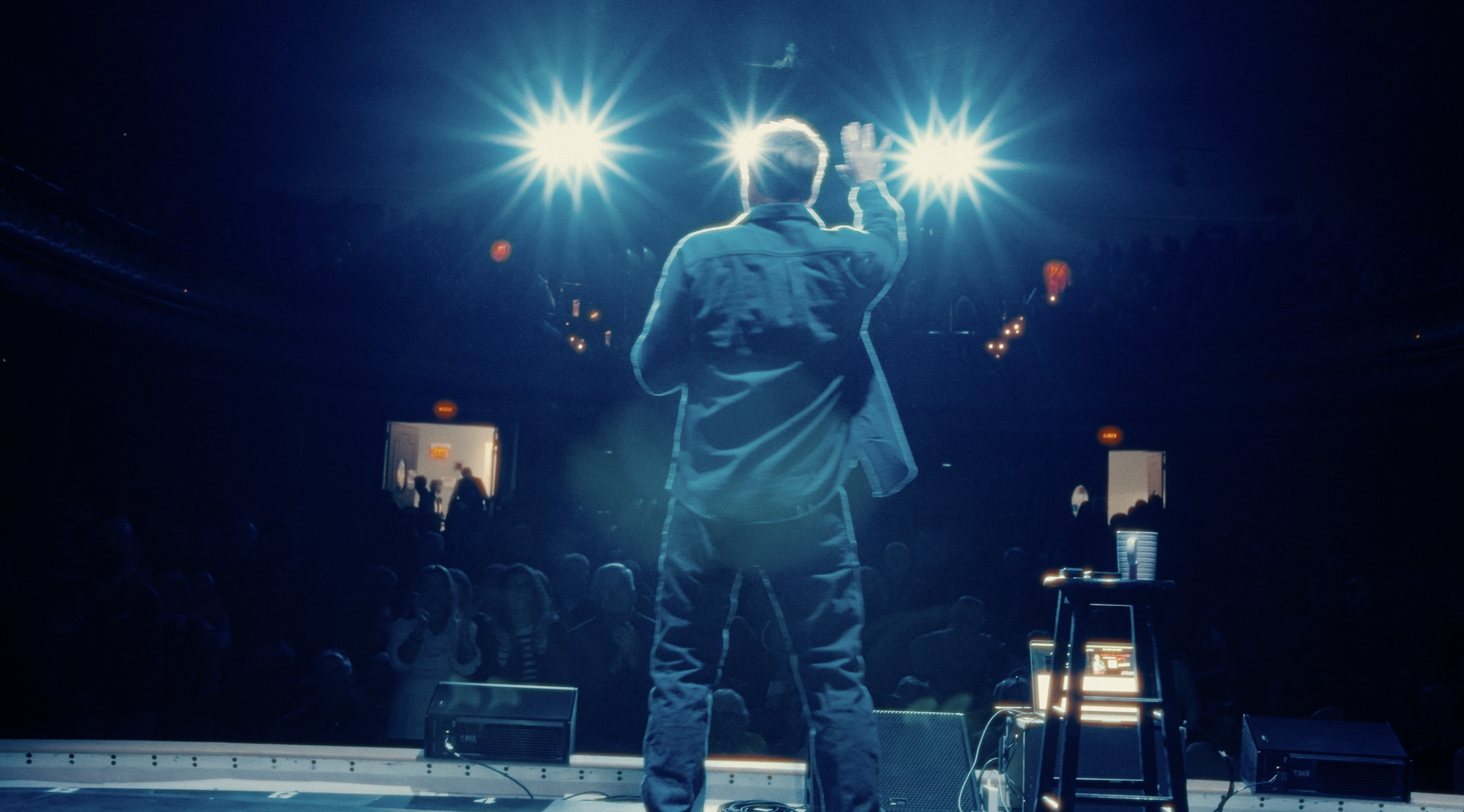 A performer on stage with his back to the camera, raising a hand, with bright stage lights shining behind him. The audience is seated in front, and some equipment can be seen on the stage.