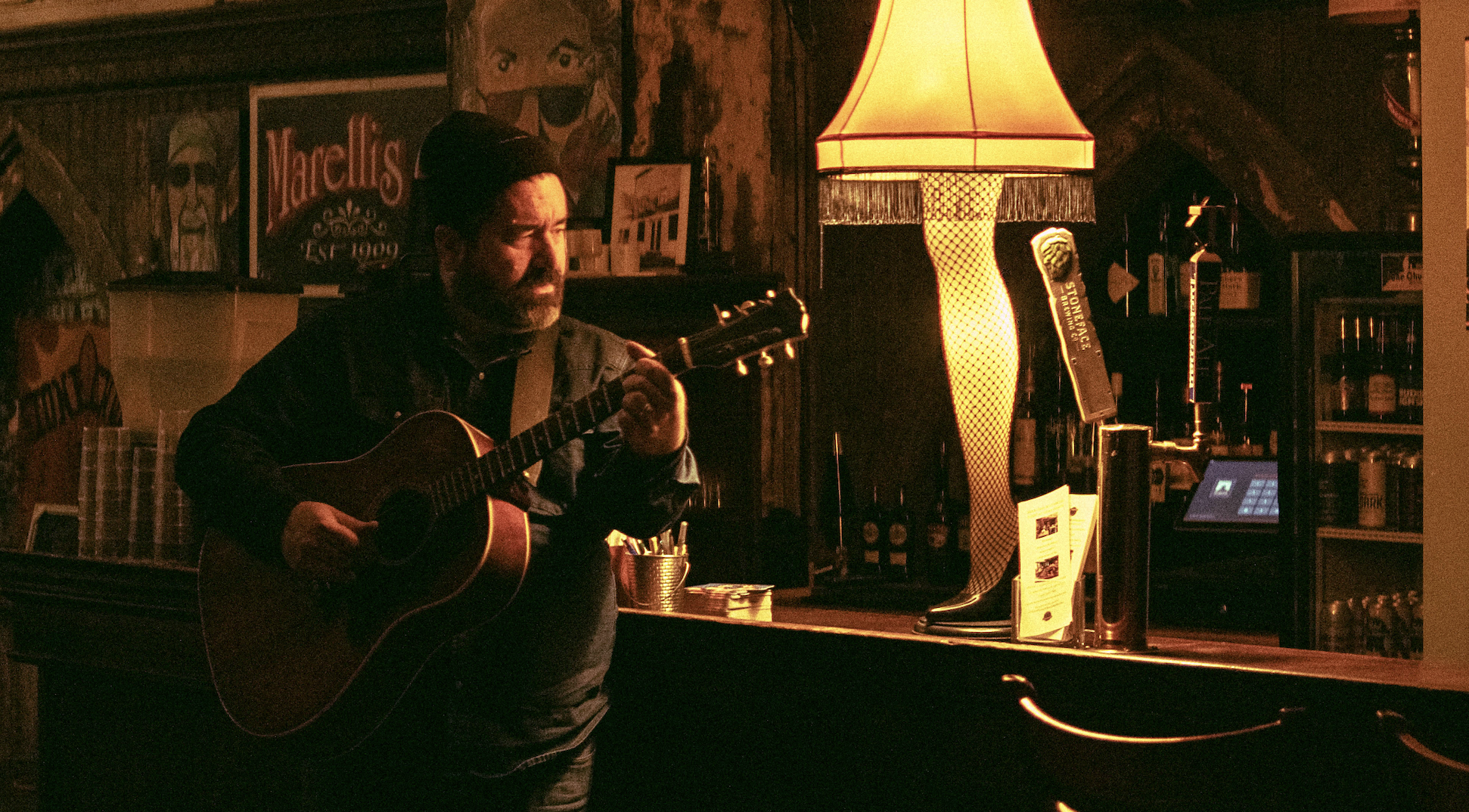 A man playing an acoustic guitar in a cozy dimly lit bar or pub. There is a large lamp with a fringed shade casting a warm glow, and various posters and pictures on the wooden walls.