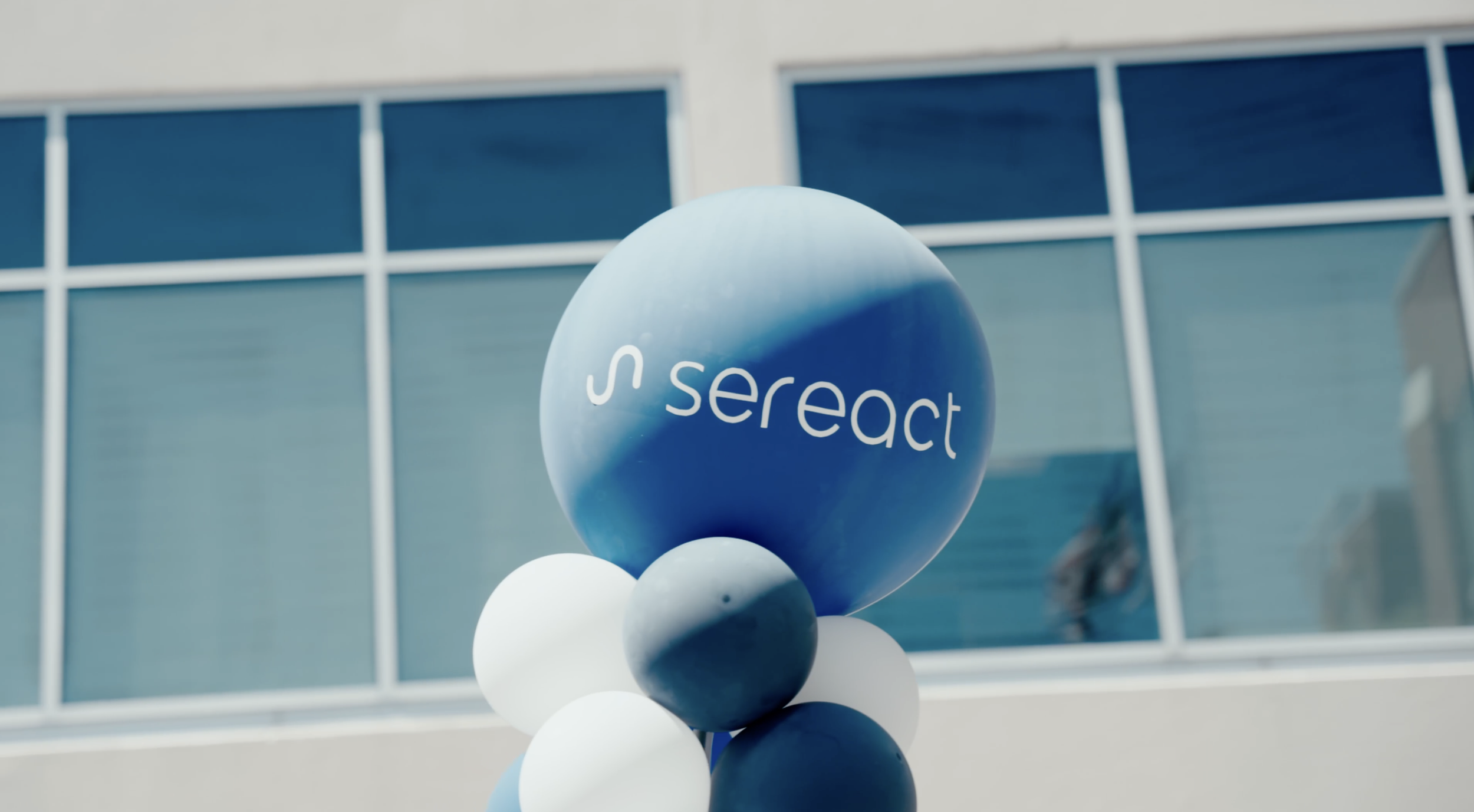 A cluster of balloons with a large blue balloon displaying the 'S react' logo, positioned in front of a modern building with reflective blue-tinted windows.
