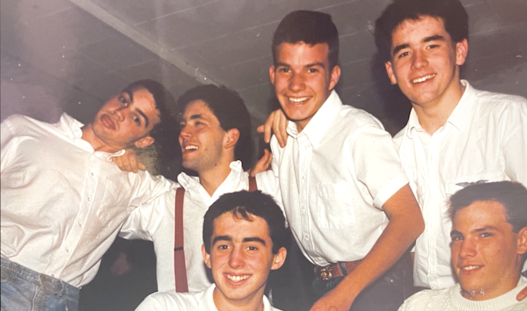 A group of six young men wearing white shirts and smiles, posing together indoors, with some showing playful and cheerful expressions.