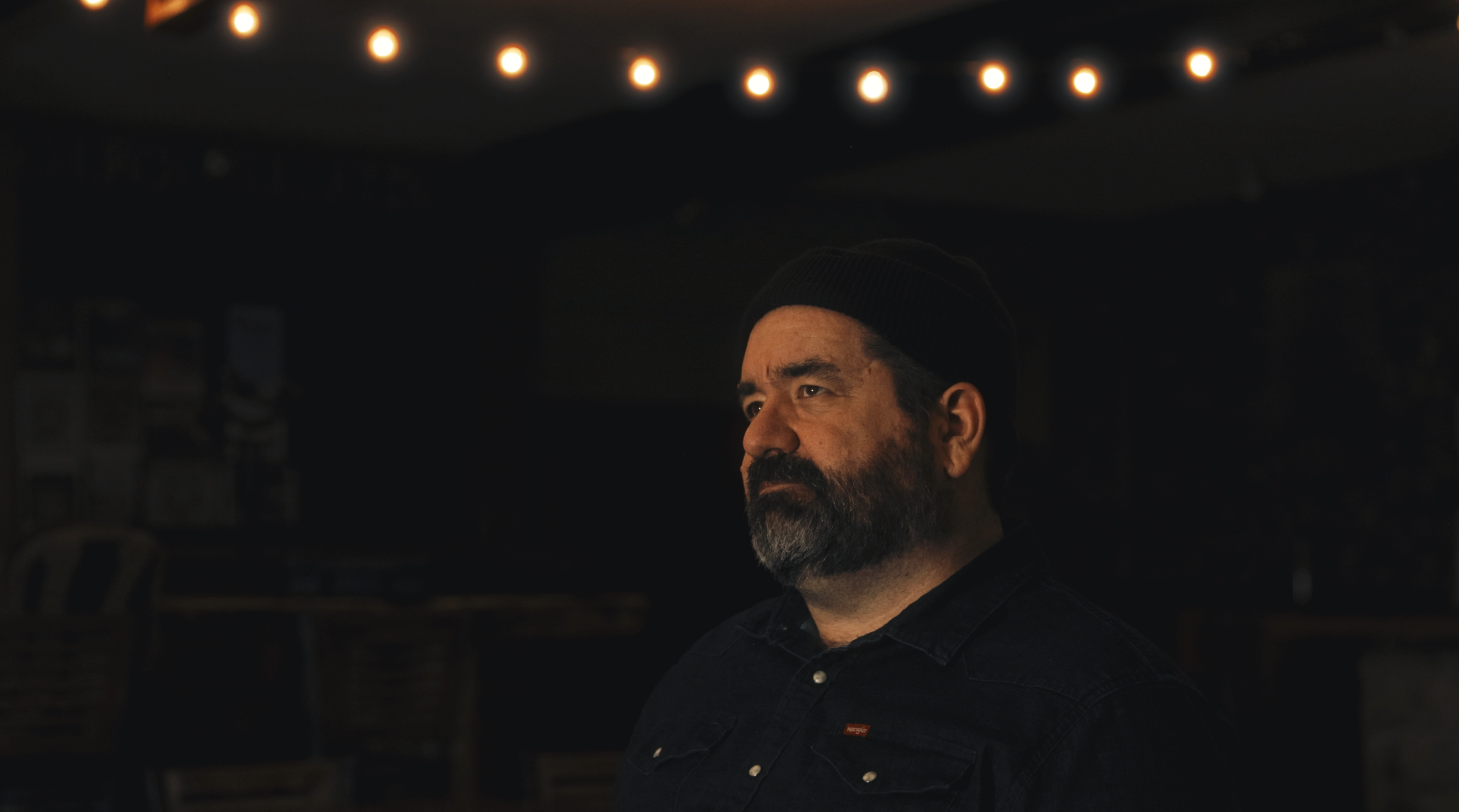 A bearded man wearing a black beanie and dark shirt in a dimly lit room with warm string lights overhead.