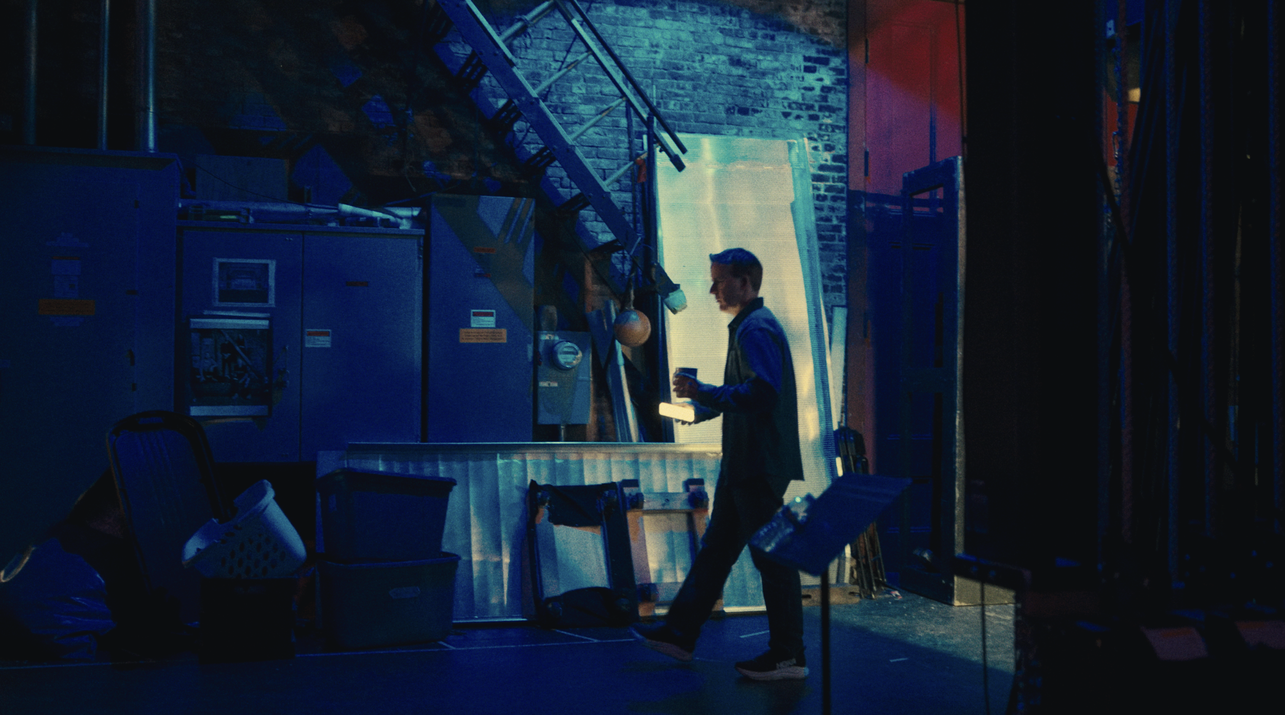 A man standing in a dimly lit industrial or backstage setting, holding a cup, with exposed brick walls, electrical panels, and metal equipment around him.
