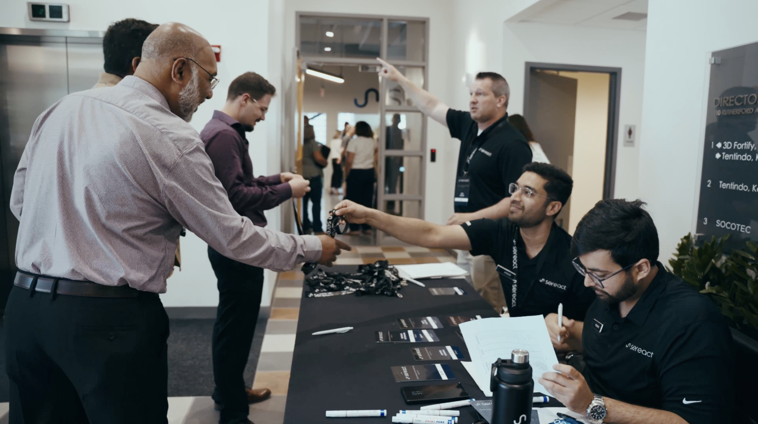 Attendees at a conference registration desk, where two staff members assist a man in handing over his registration bag, with other people in the background and informational signage visible.