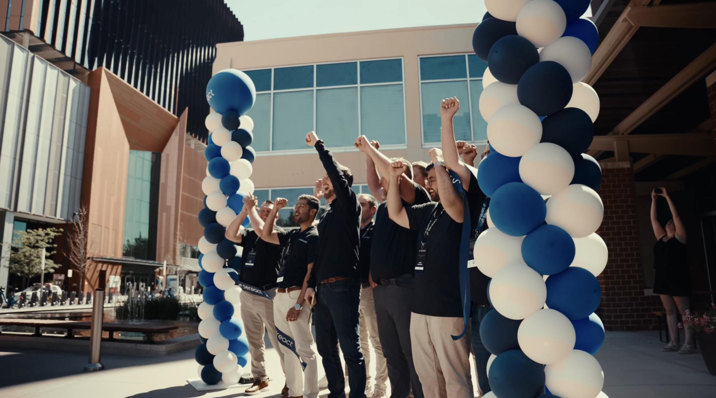 Group of people celebrating under a balloon arch outside a modern building, raising their fists, smiling, and cheering.