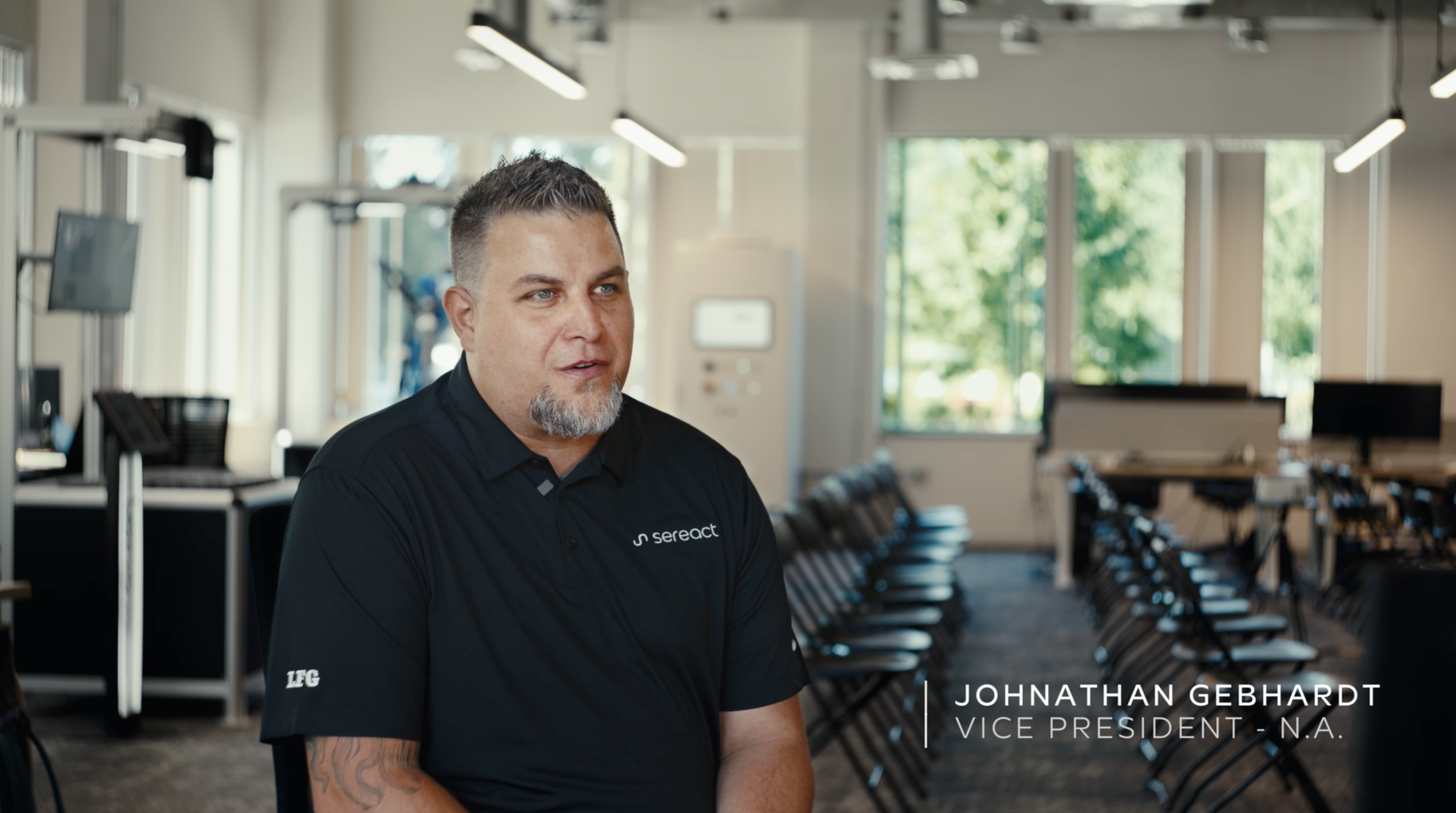 A man with short hair and a beard wearing a black polo shirt with a logo, sitting in a modern conference room with large windows, chairs, and screens. The text overlay identifies him as Johnathan Gebhardt, Vice President of N.A.