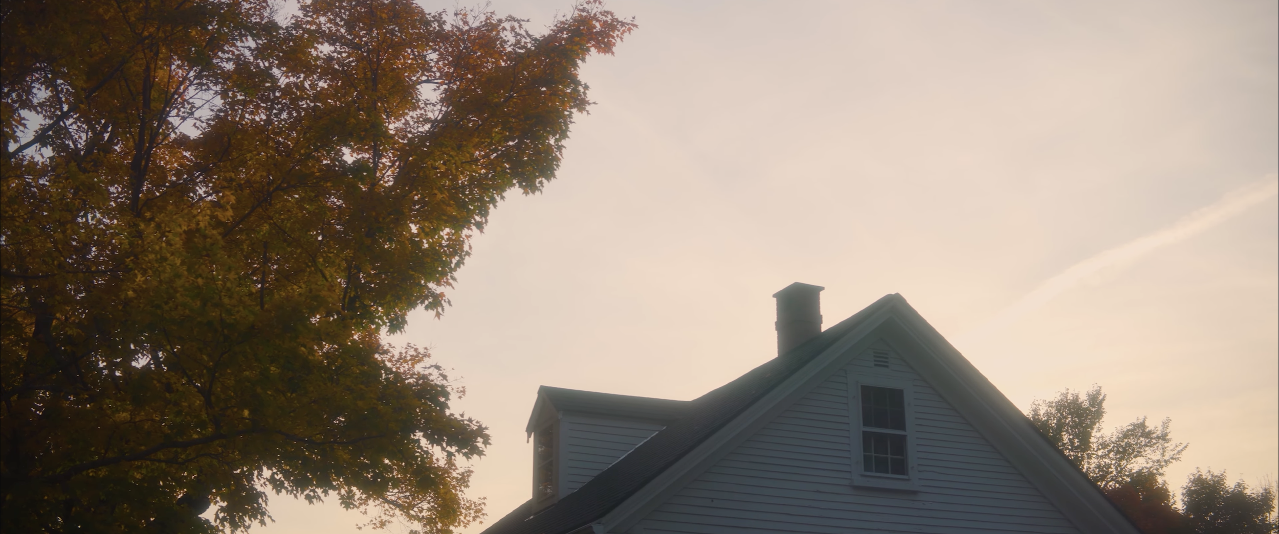 A house with white siding and a chimney, partially obscured by trees with autumn-colored leaves, under a pale sky at sunset.