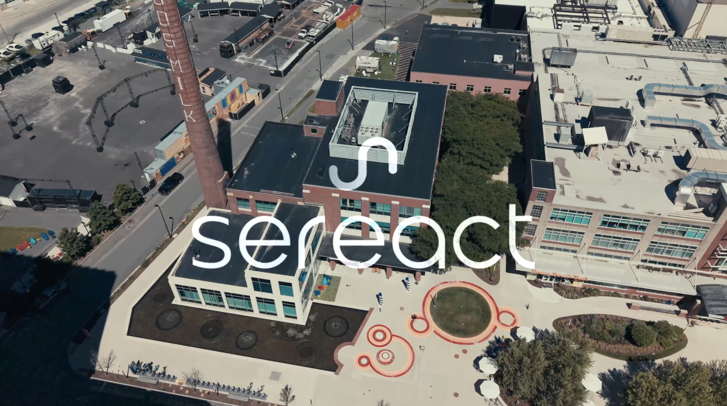 Aerial view of a building with a rooftop HVAC system, adjacent to a playground with circular patterns, parking lot and surrounding cityscape.