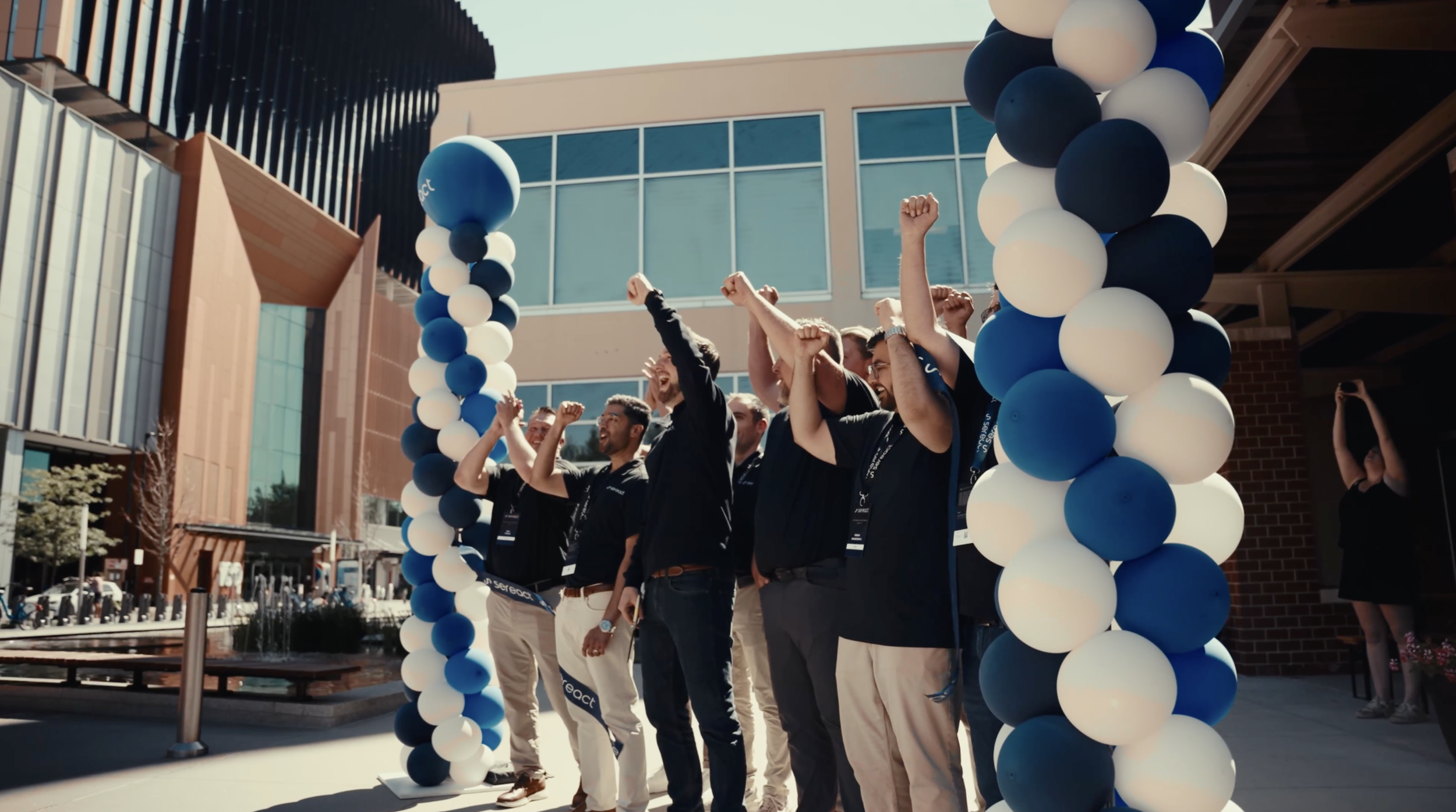 A group of people standing under a balloon arch, raising their hands in celebration outside a modern building.