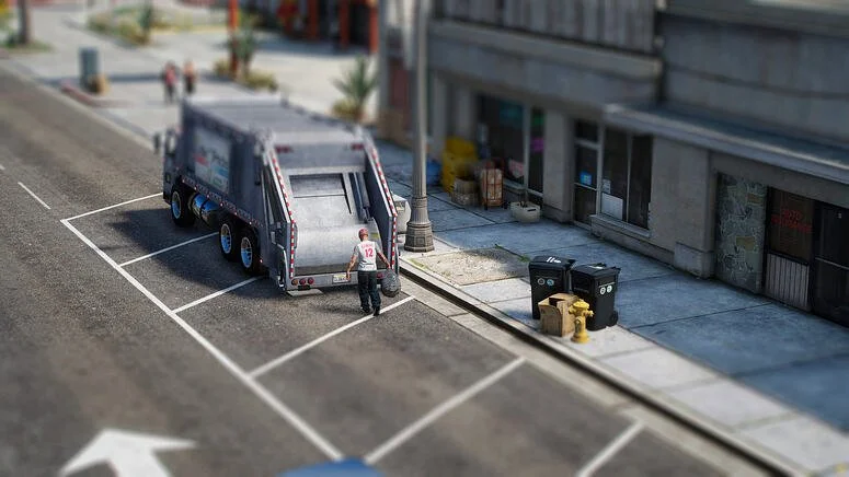 A sanitation worker unloading trash from a black garbage truck on a city street, with side of building, sidewalk, trash cans, and fire hydrant visible.