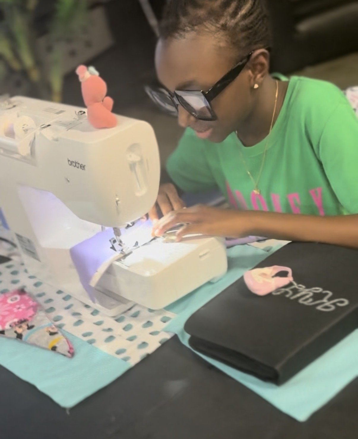 A young woman sewing on a white Brother sewing machine, with a pink plush toy on top, at a table covered with a turquoise tablecloth. She wears glasses and a green shirt.