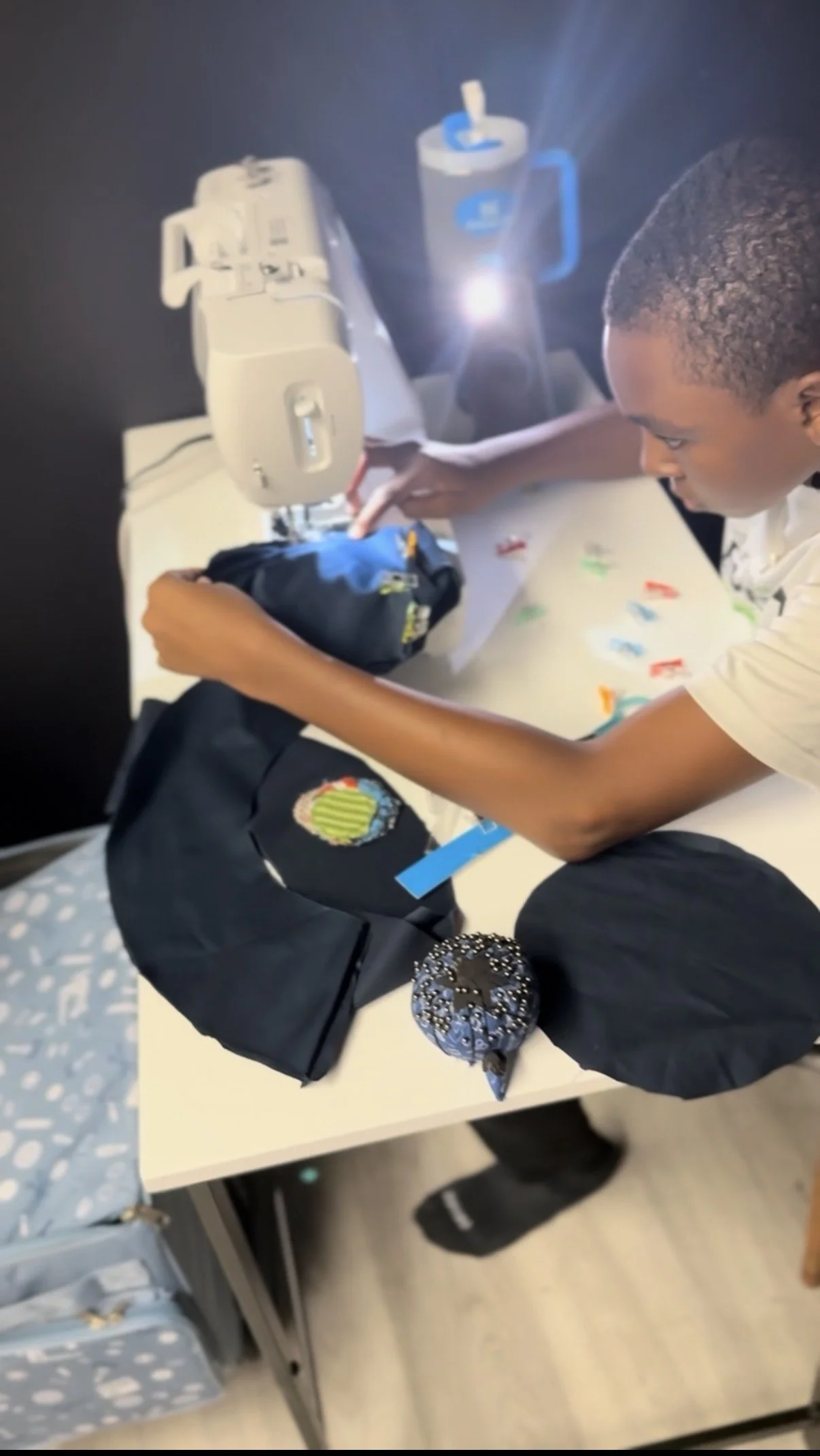 A boy operating a sewing machine at a sewing table with a black backpack, a decorative bag, and a container nearby at Andreya Lovelace's children's sewing summer camp.