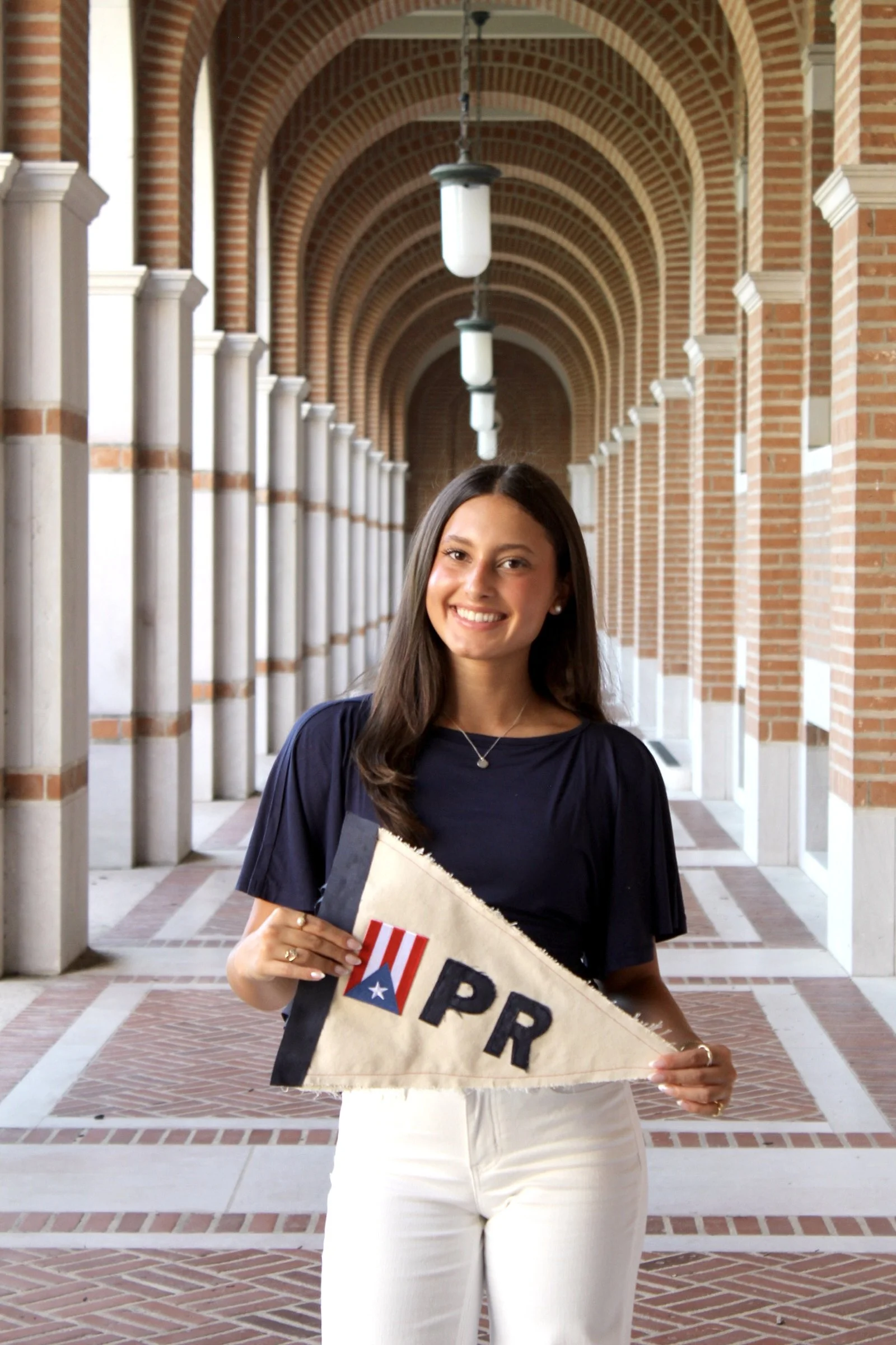 A young woman in a navy blue top and white pants holding a surfboard with an American flag design and a Texas flag, standing in an arched brick corridor with hanging lights.