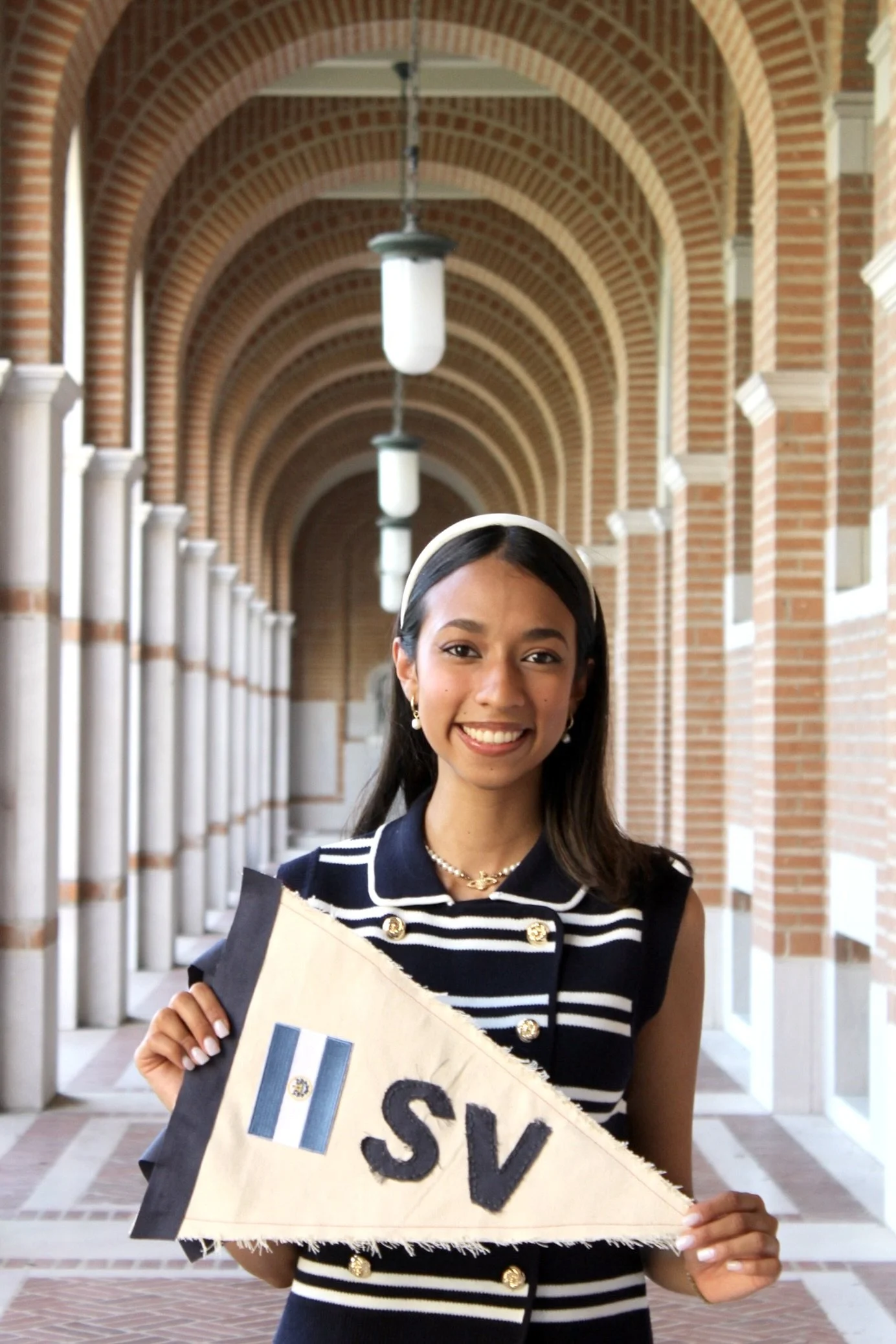 A young woman smiling, holding a handmade sign with a small flag sticker and the word 'SV' on it, standing in a brick arcway corridor with hanging lights.