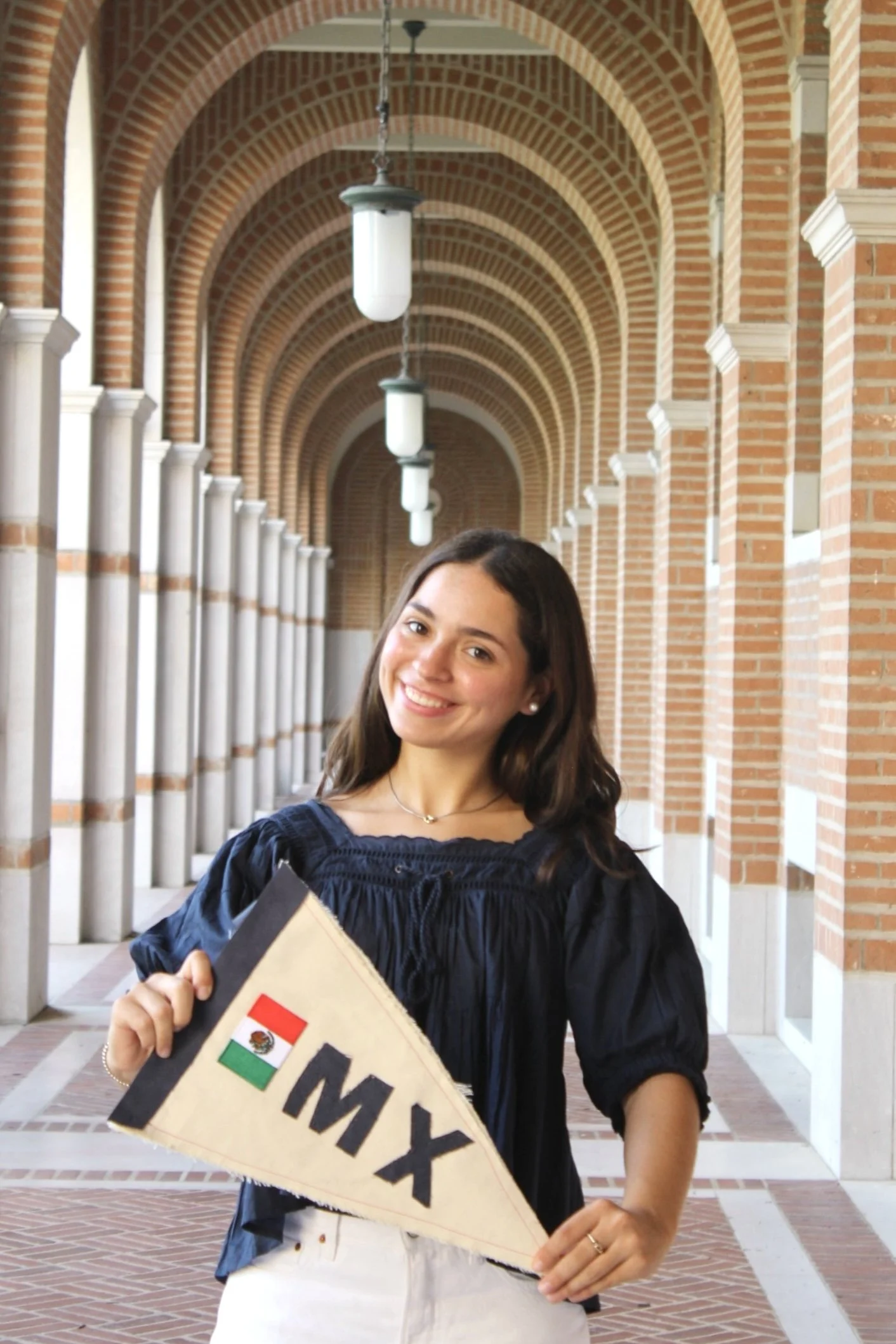 A smiling young woman in a graduation gown holding a sign with the Mexican flag in an arched brick corridor.