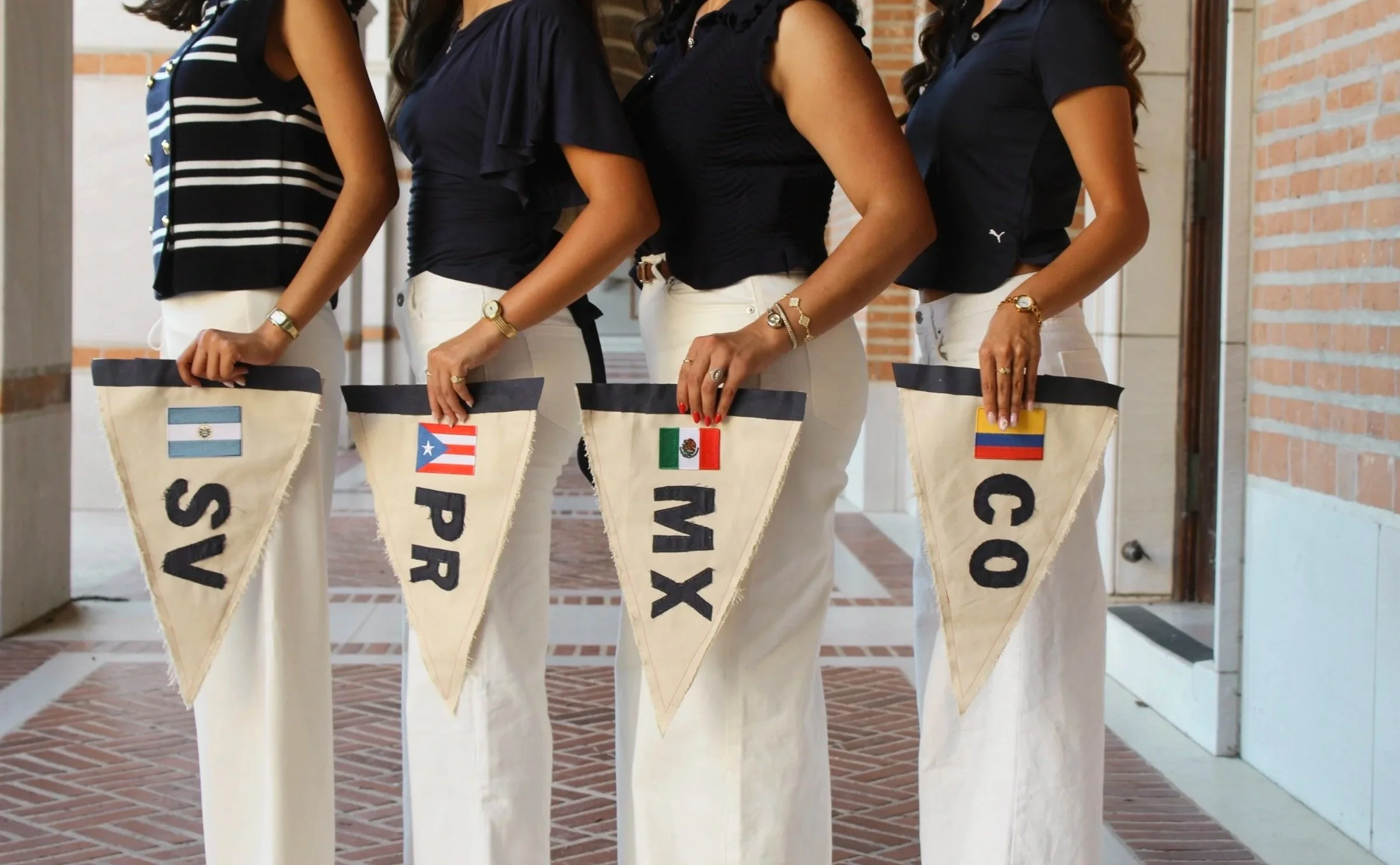 Four women standing in a line, each holding a felt pennant with their country's flag and some text, dressed in white pants and navy tops, in an indoor setting with brick walls.