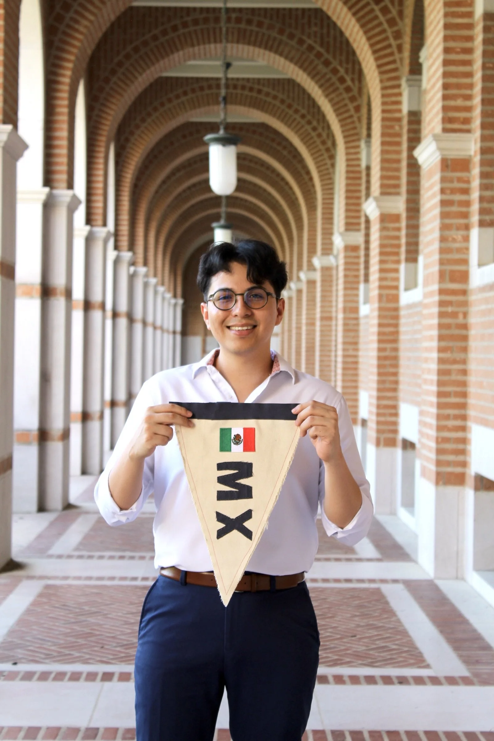 Young man in white shirt and dark pants smiling, holding a triangular banner with the Mexican flag and the word "MEX" on it, standing in an arched brick corridor.