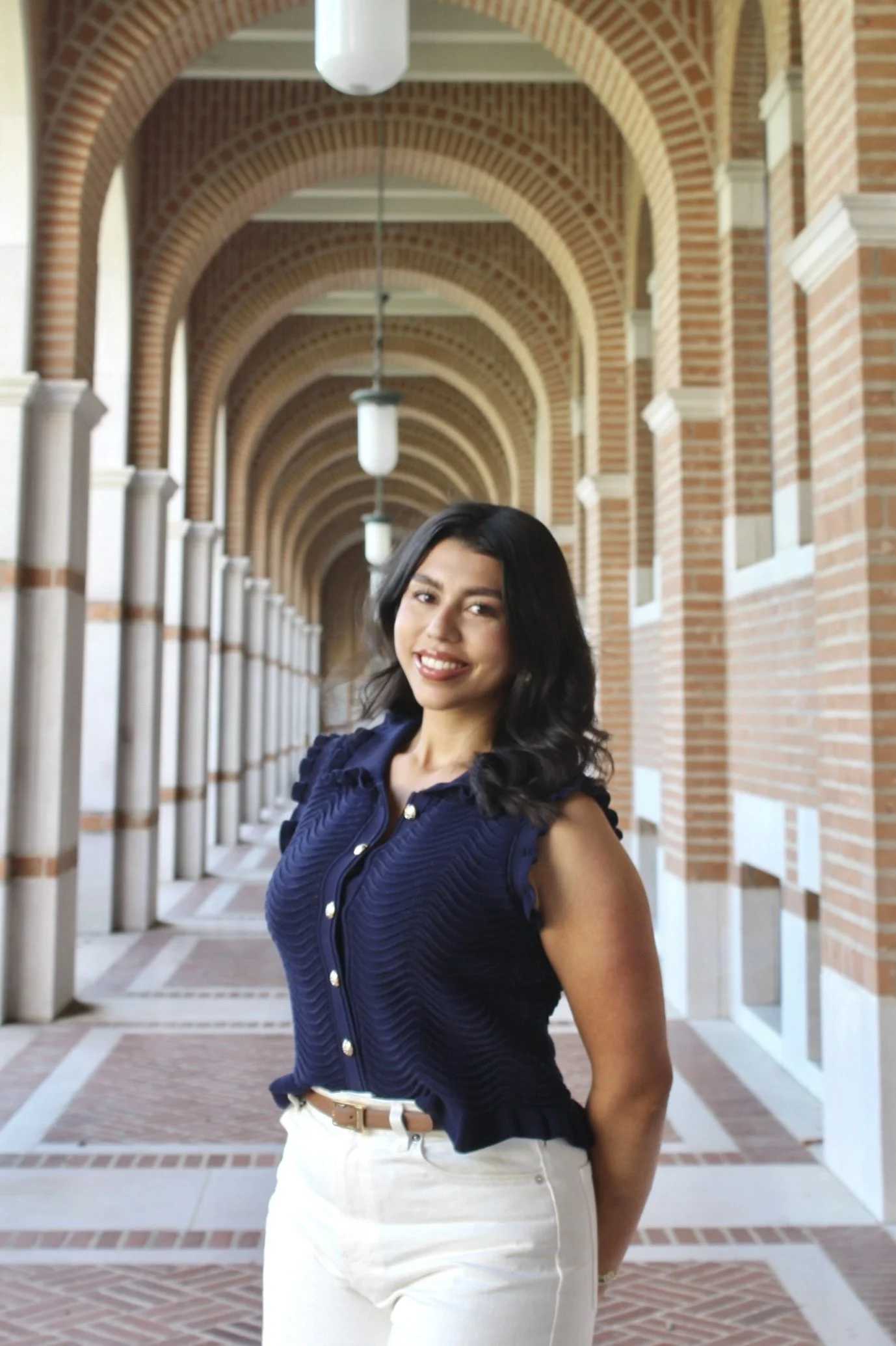 A woman with black hair smiling and wearing a navy blue sleeveless top and white pants, standing in a brick arched walkway with hanging lights.