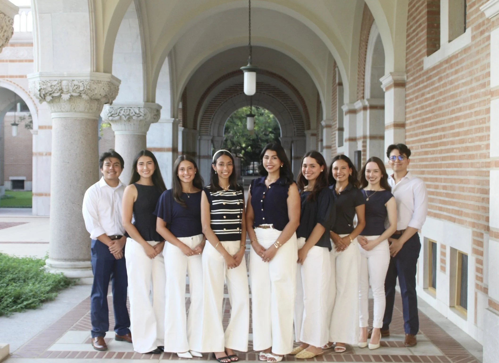 Group of ten young adults standing outside a building with brick walls, arches, and columns, smiling at the camera.