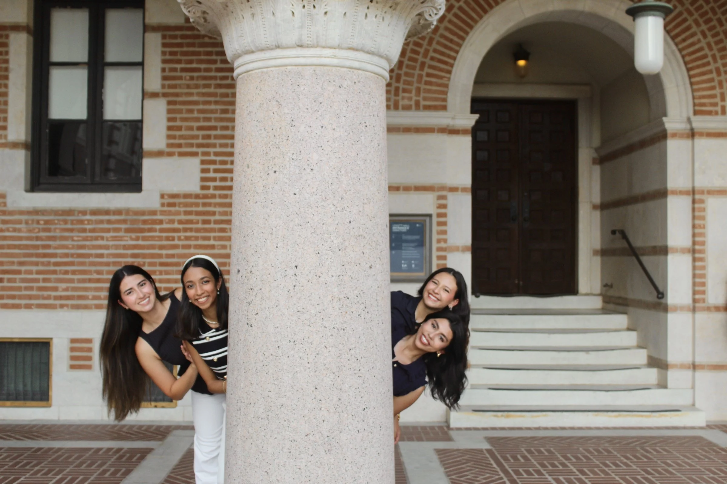 Group of five women, smiling and leaning at an angle, posing behind a large stone column in front of a brick building with stairs.