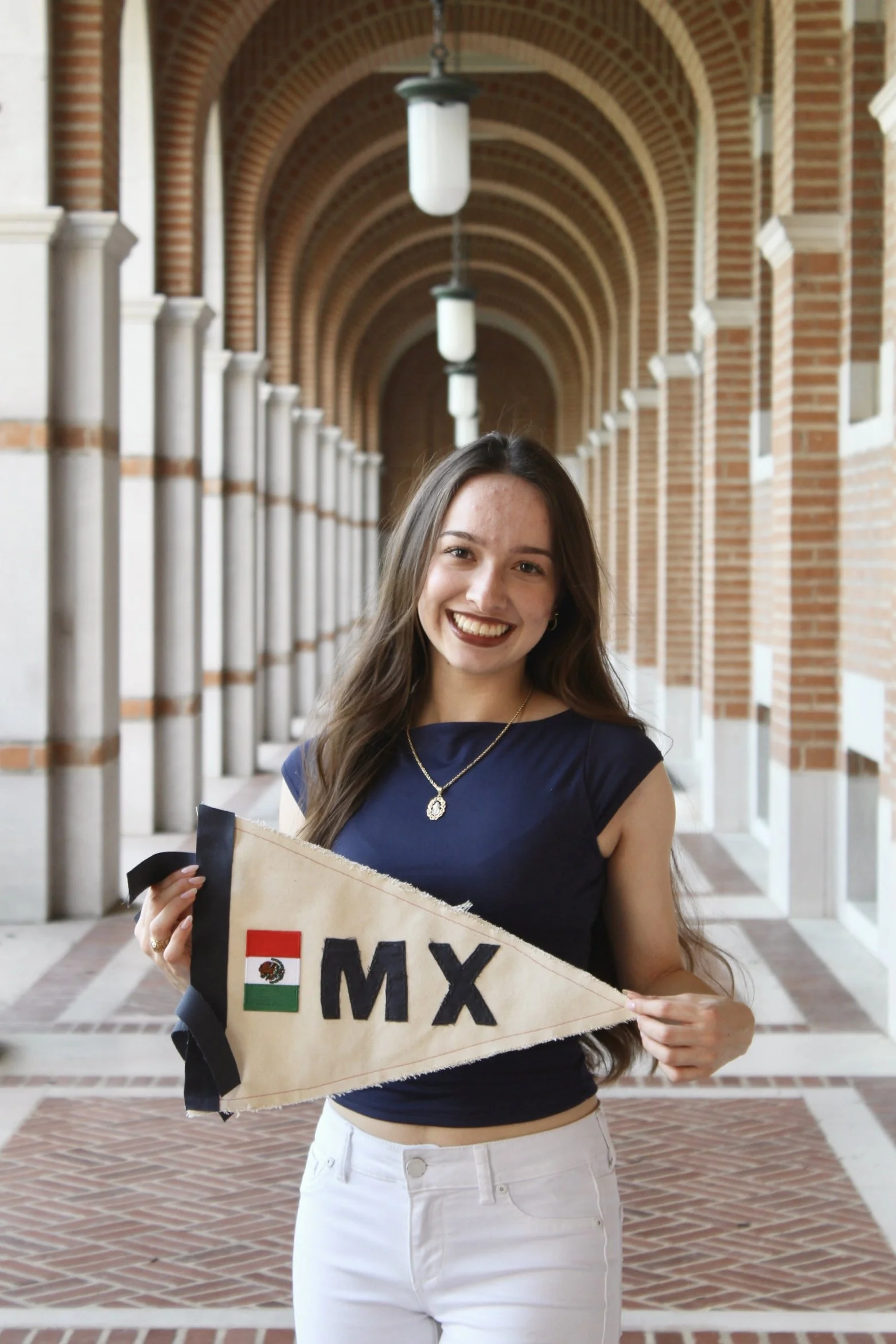A woman with long brown hair smiling, holding a Mexican flag banner with the text 'MEX' and an embroidered Mexican flag, standing in a brick corridor with arches and hanging lights.