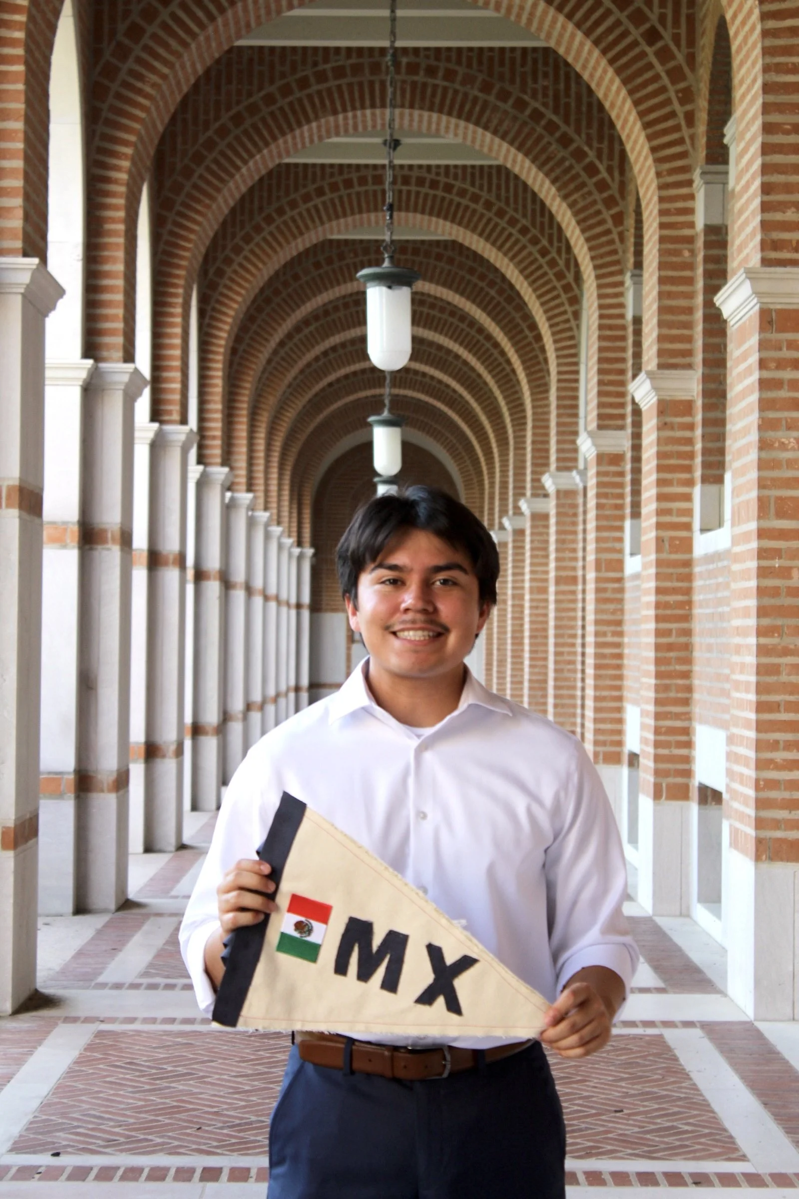 Young man in white shirt holding a sign with the letters 'MX' and an Indian flag, standing in a corridor with brick arches and hanging lights.