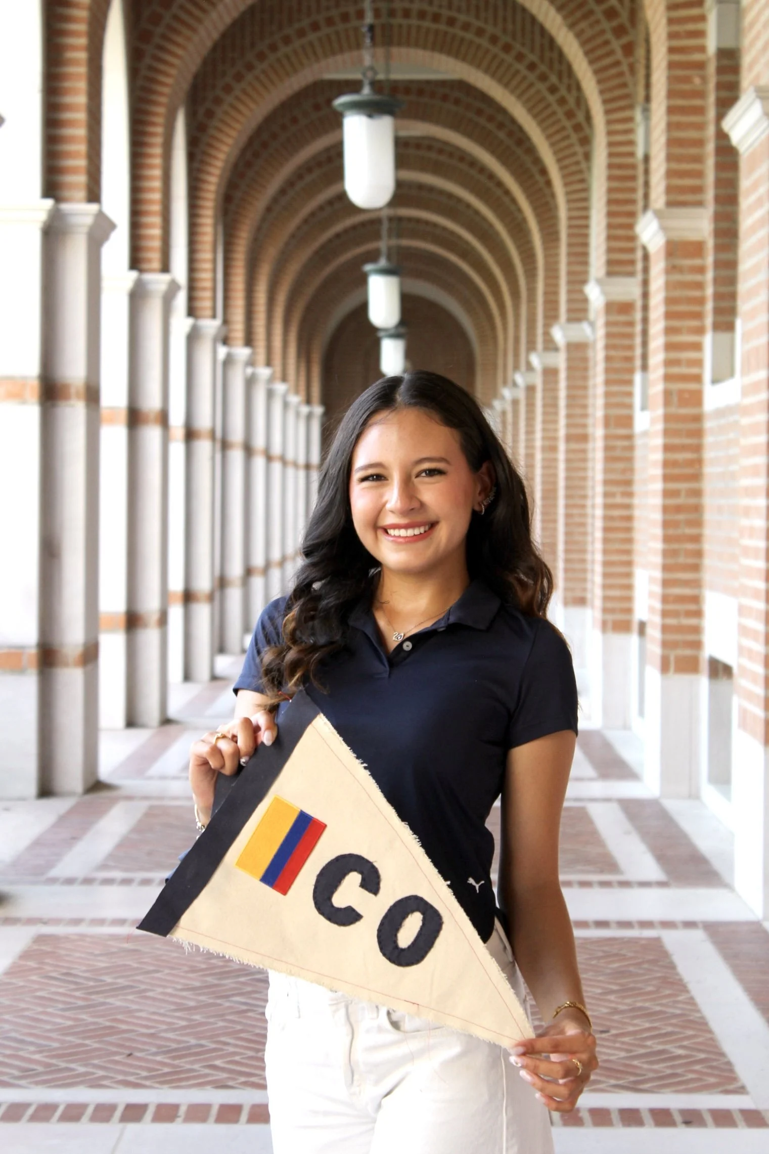 A young woman with long dark hair and a smile, holding a handmade graduation cap with a Colombian flag and the word 'SOCIO' written on it, standing under a brick archway with hanging lights.