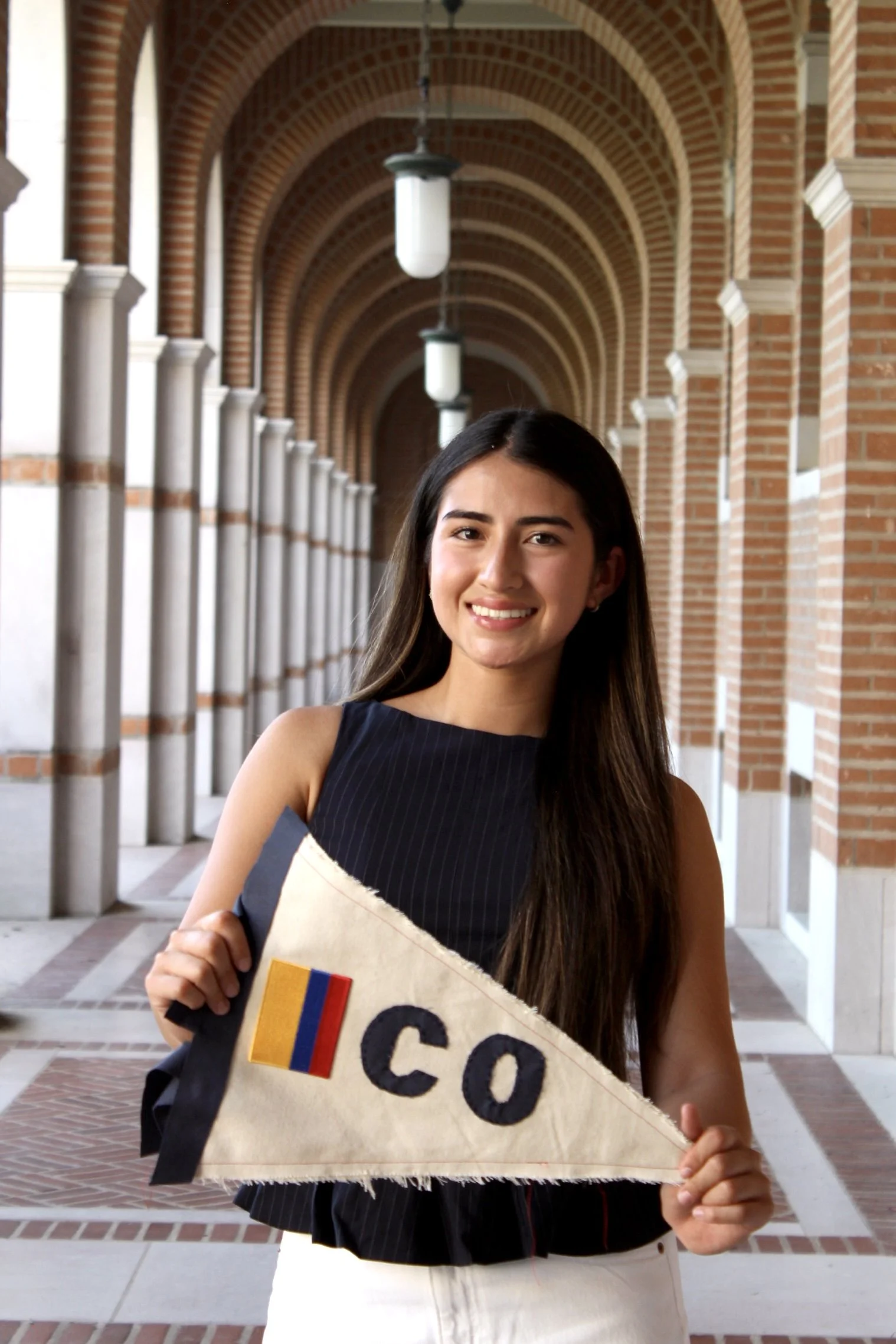 A young woman standing in an arched brick corridor, smiling while holding a graduation cap with a colorful patch and a diploma.