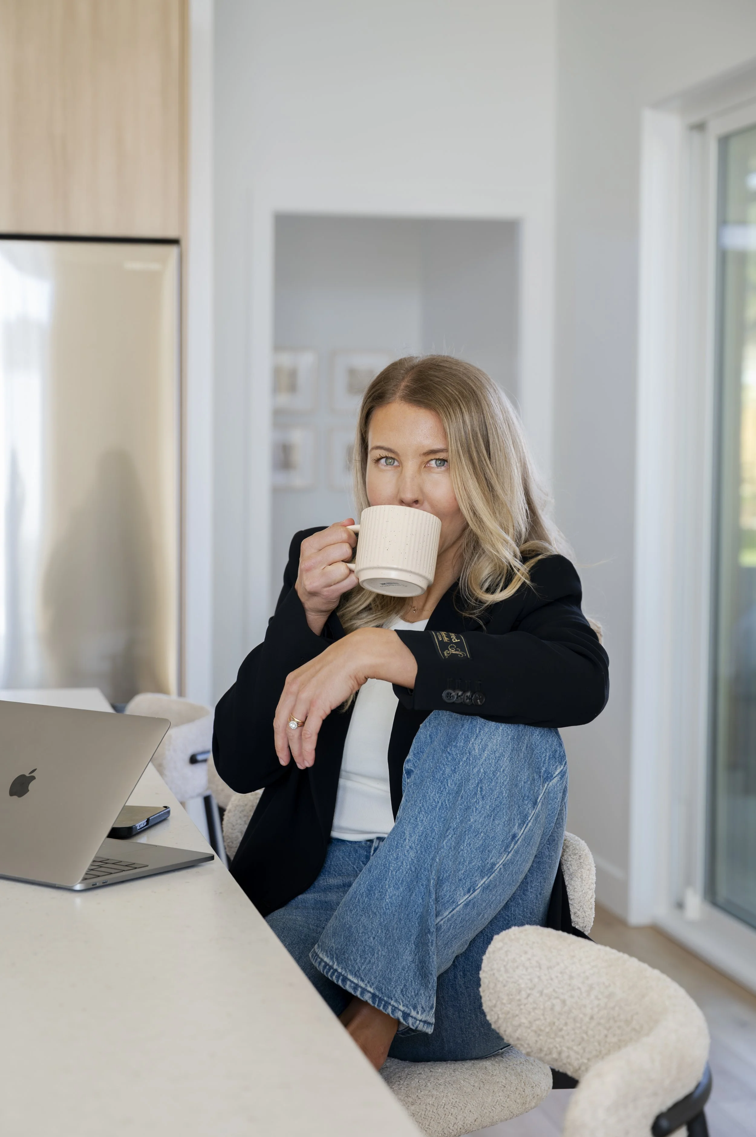 Woman sitting at a desk, drinking from a mug, with a laptop open in front of her. She is wearing a black blazer and jeans, with a teddy bear on her lap. The room appears bright with natural light coming from large windows.