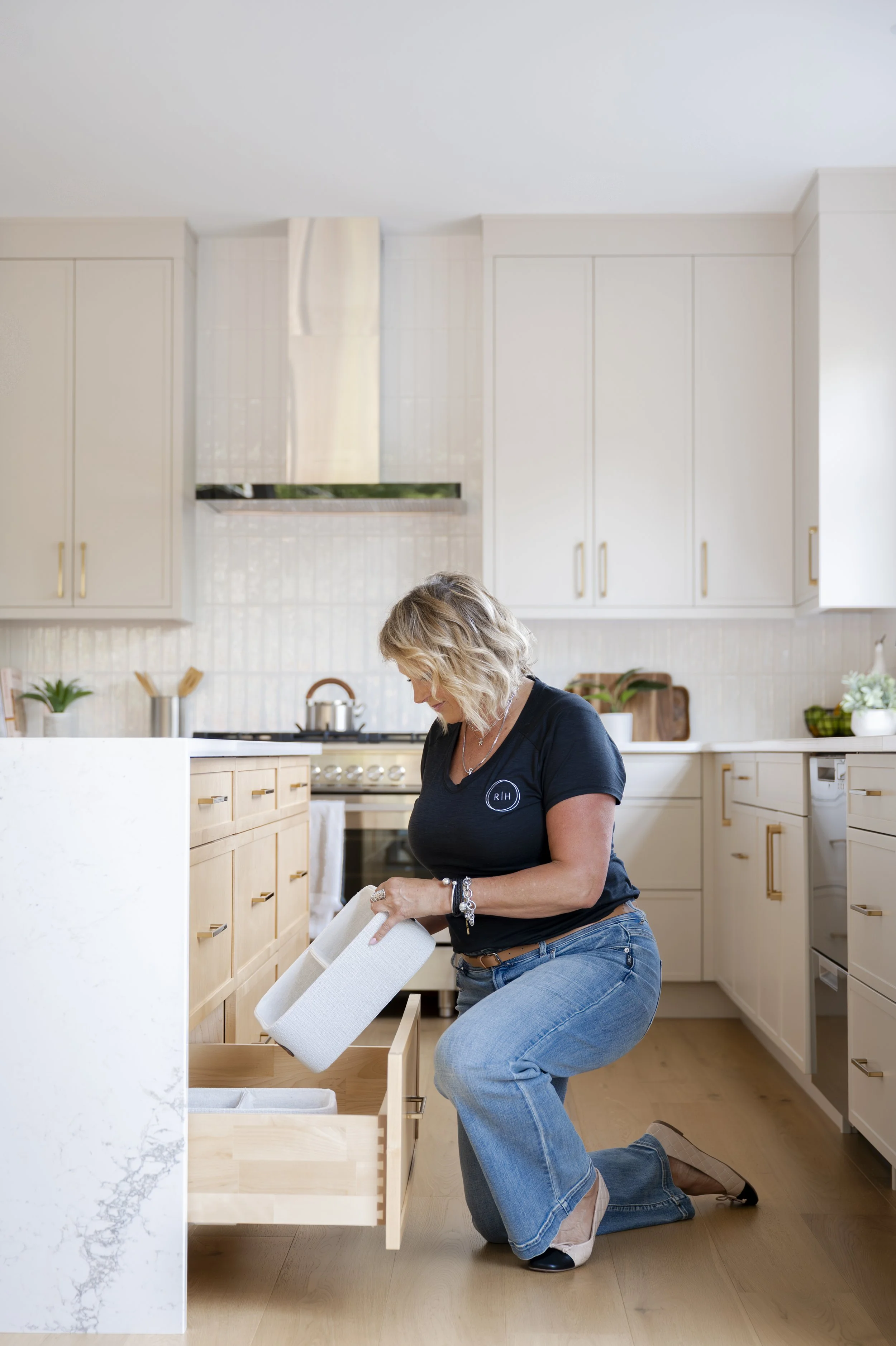 Woman kneeling on the kitchen floor opening a drawer in light wood cabinets, with a white countertop with a marble pattern in the foreground.