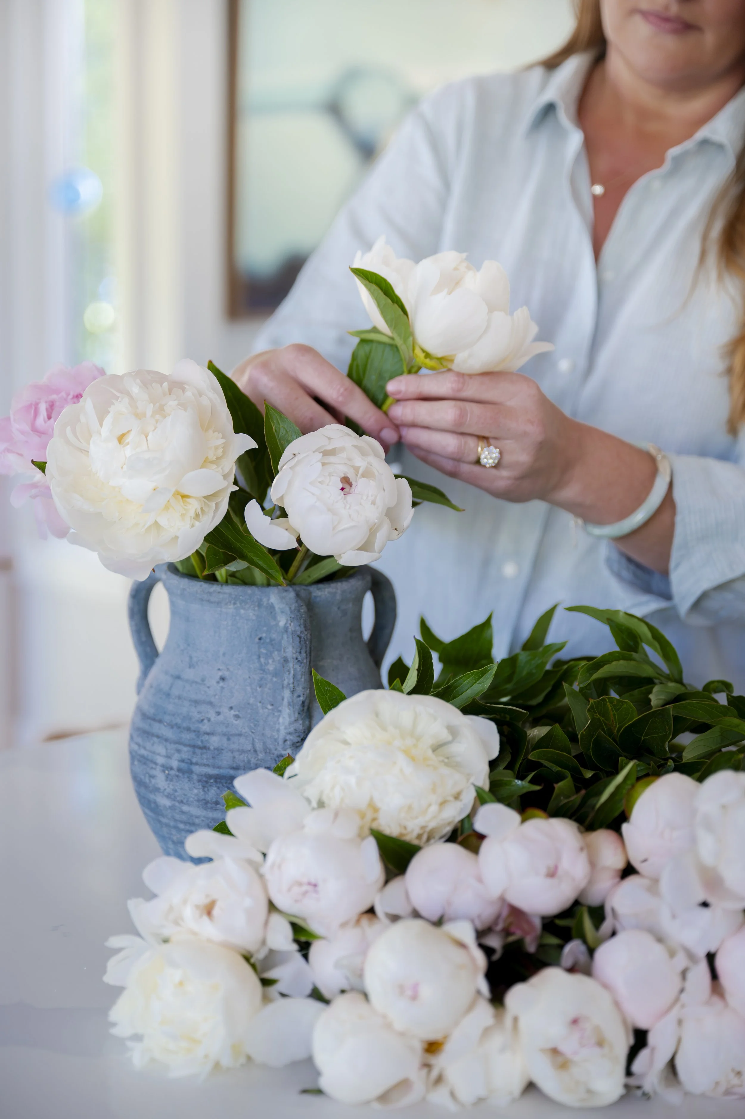 A woman arranging white and pink peonies in a blue ceramic vase, surrounded by various greenery, indoors.