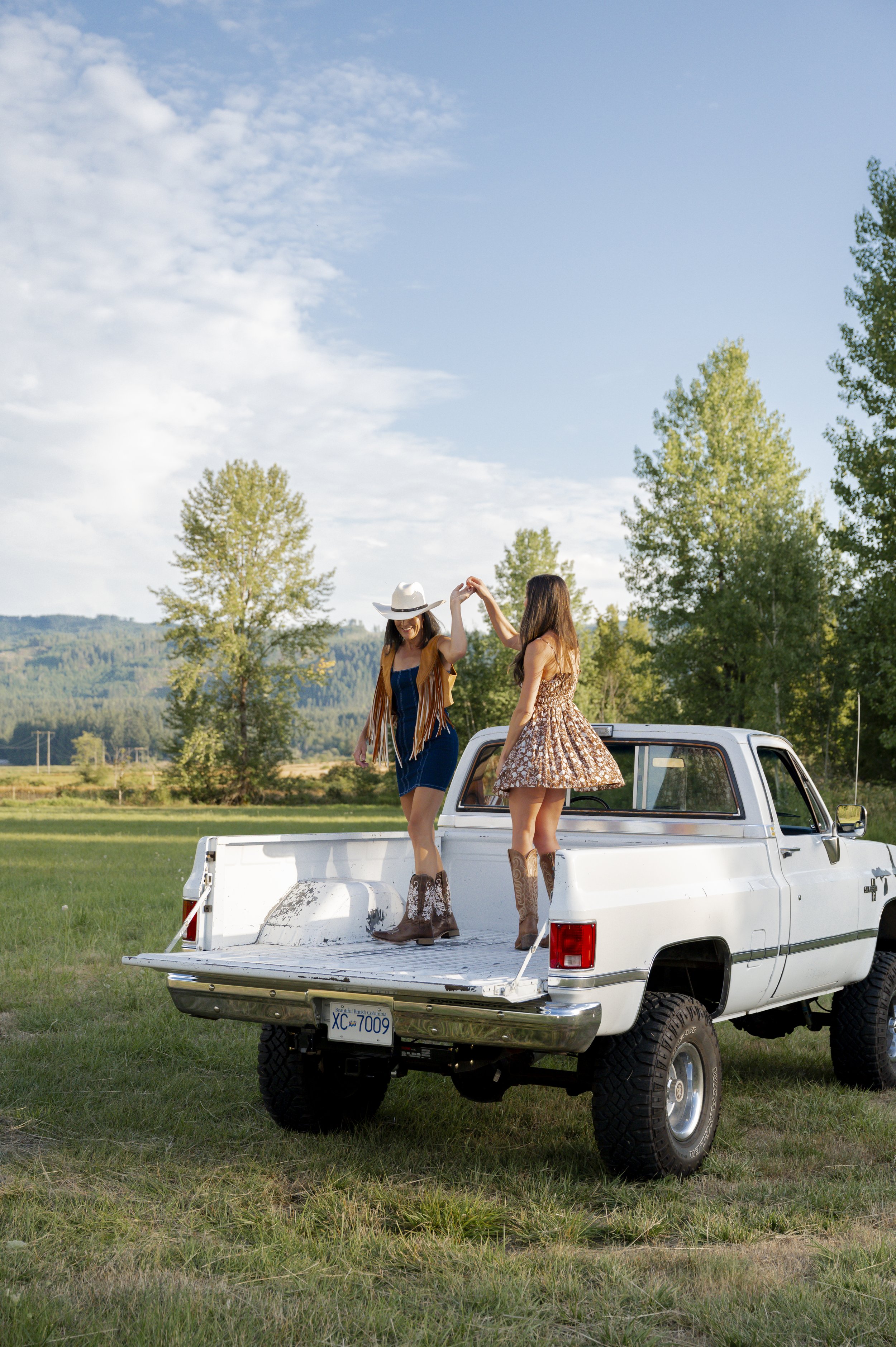 Two women in cowboy boots and dresses dancing on the bed of a white pickup truck in a grassy field. One woman wears a wide-brimmed hat, and both are holding hands, with a backdrop of trees and a blue sky with clouds.