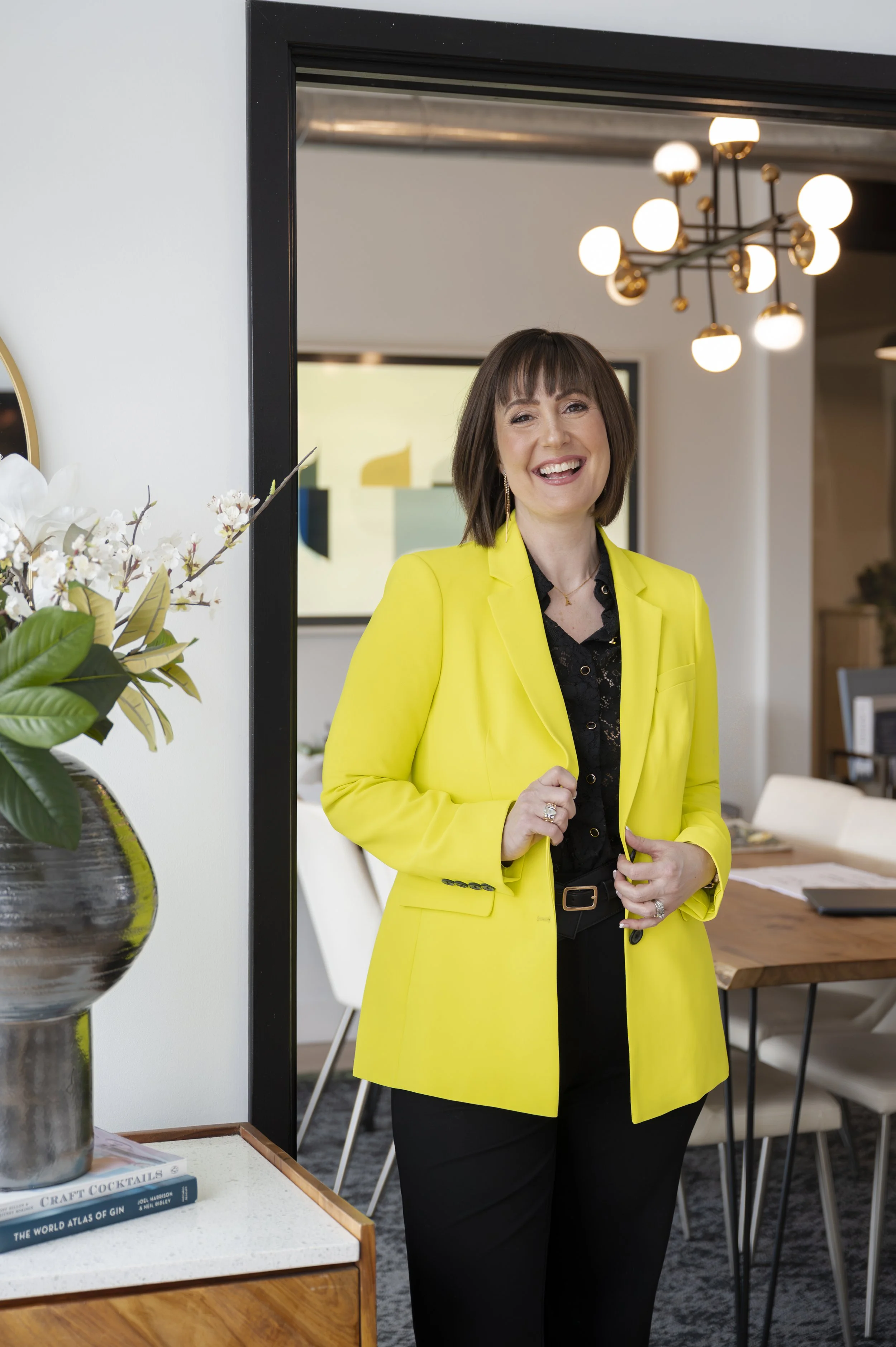 A woman with shoulder-length brown hair, smiling and squinting slightly, wearing a bright yellow blazer over a black top and black pants, standing inside a modern office space.