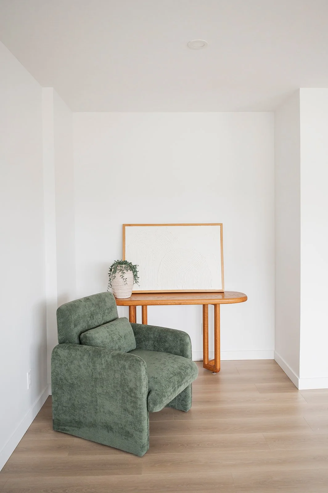 A green plush armchair with a matching pillow, a wooden side table with a white framed abstract art piece and a potted plant on top, against white walls and light wood flooring.
